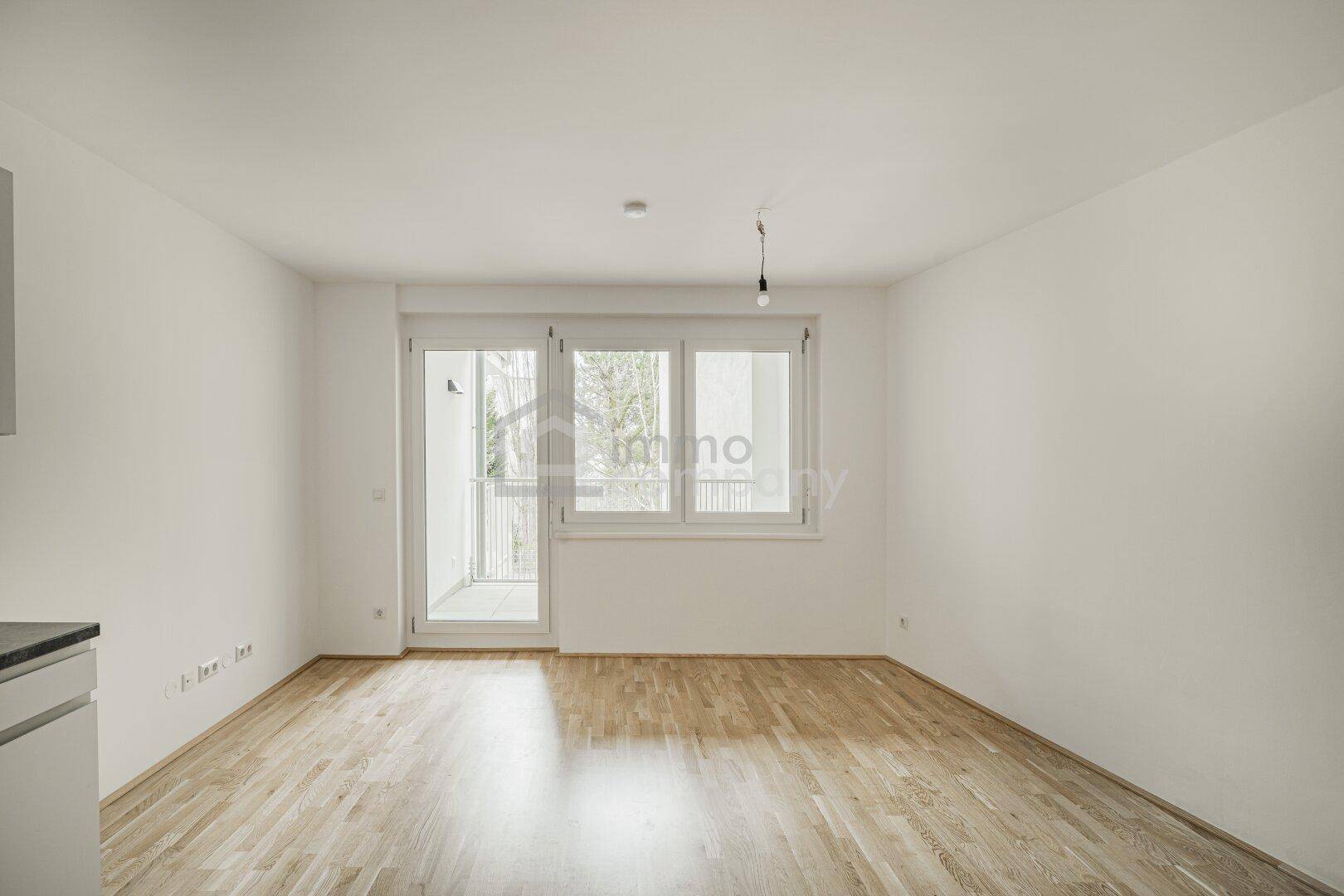 Leerer Wohnraum mit Eichenparkettboden und großen Fenstern zum Balkon.