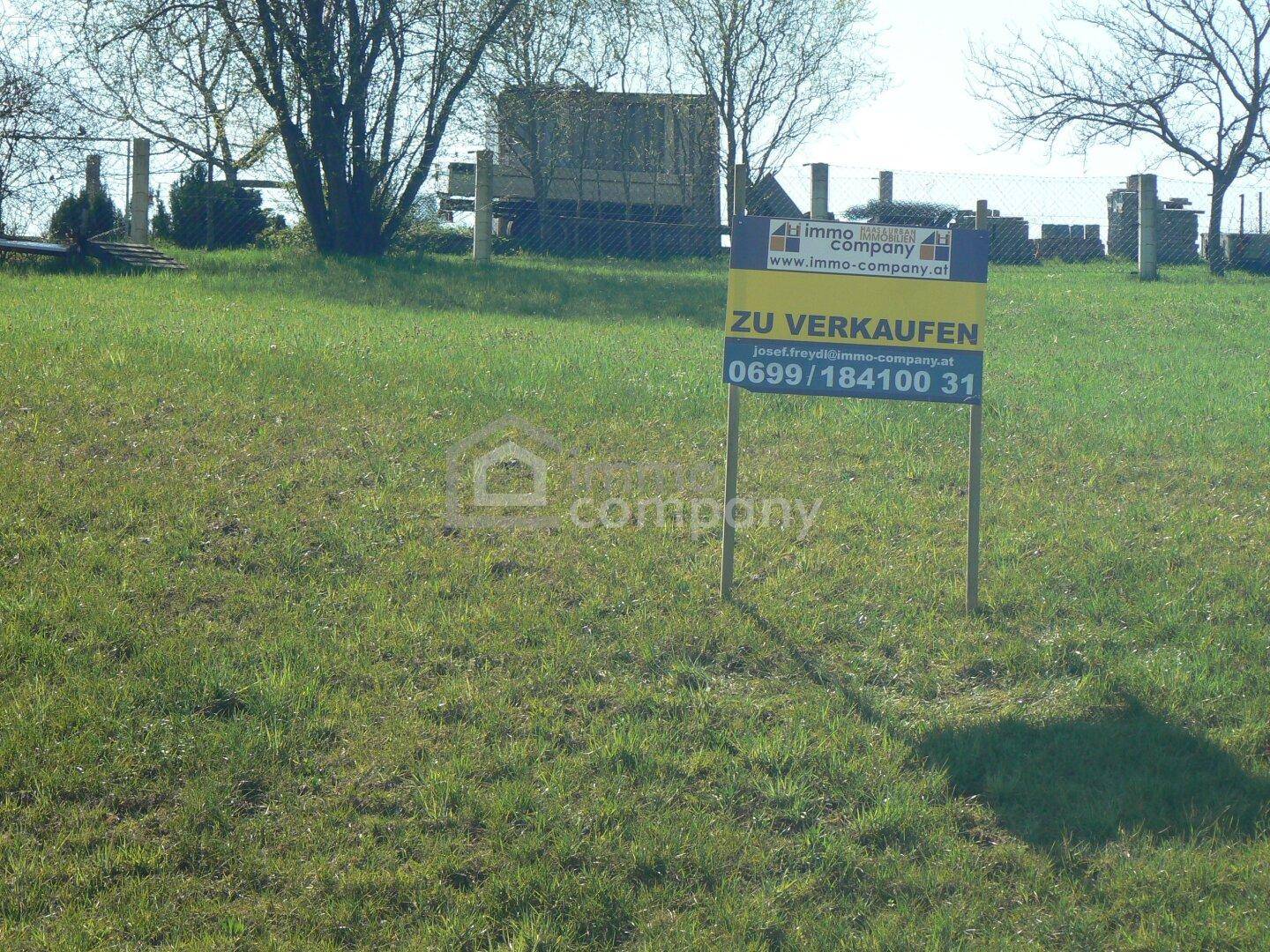 Grünes Grundstück mit einem 'Zu Verkaufen'-Schild, umgeben von ländlicher Vegetation.
