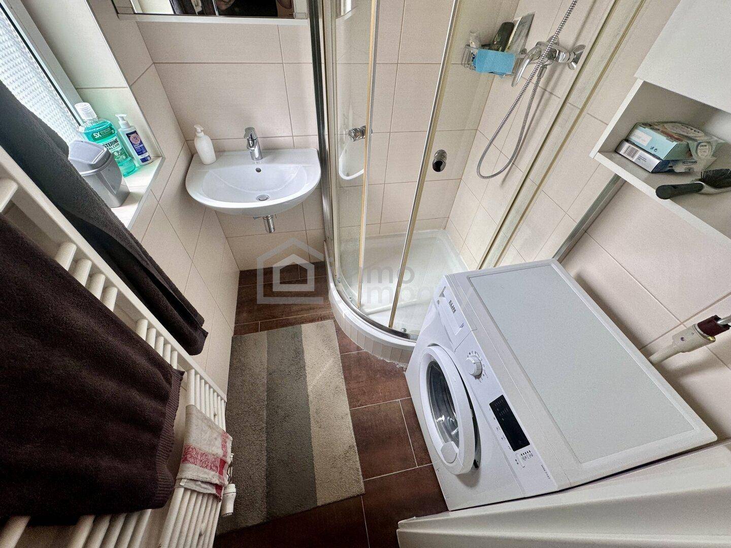 Kleines Badezimmer mit Dusche, Waschbecken, Fenster und integrierter Waschmaschine.