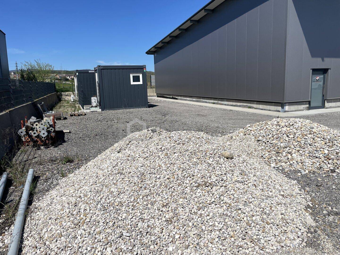 Außenansicht mit einem großen Kieshaufen, einem modernen Industriebau und kleineren Containern unter blauem Himmel.