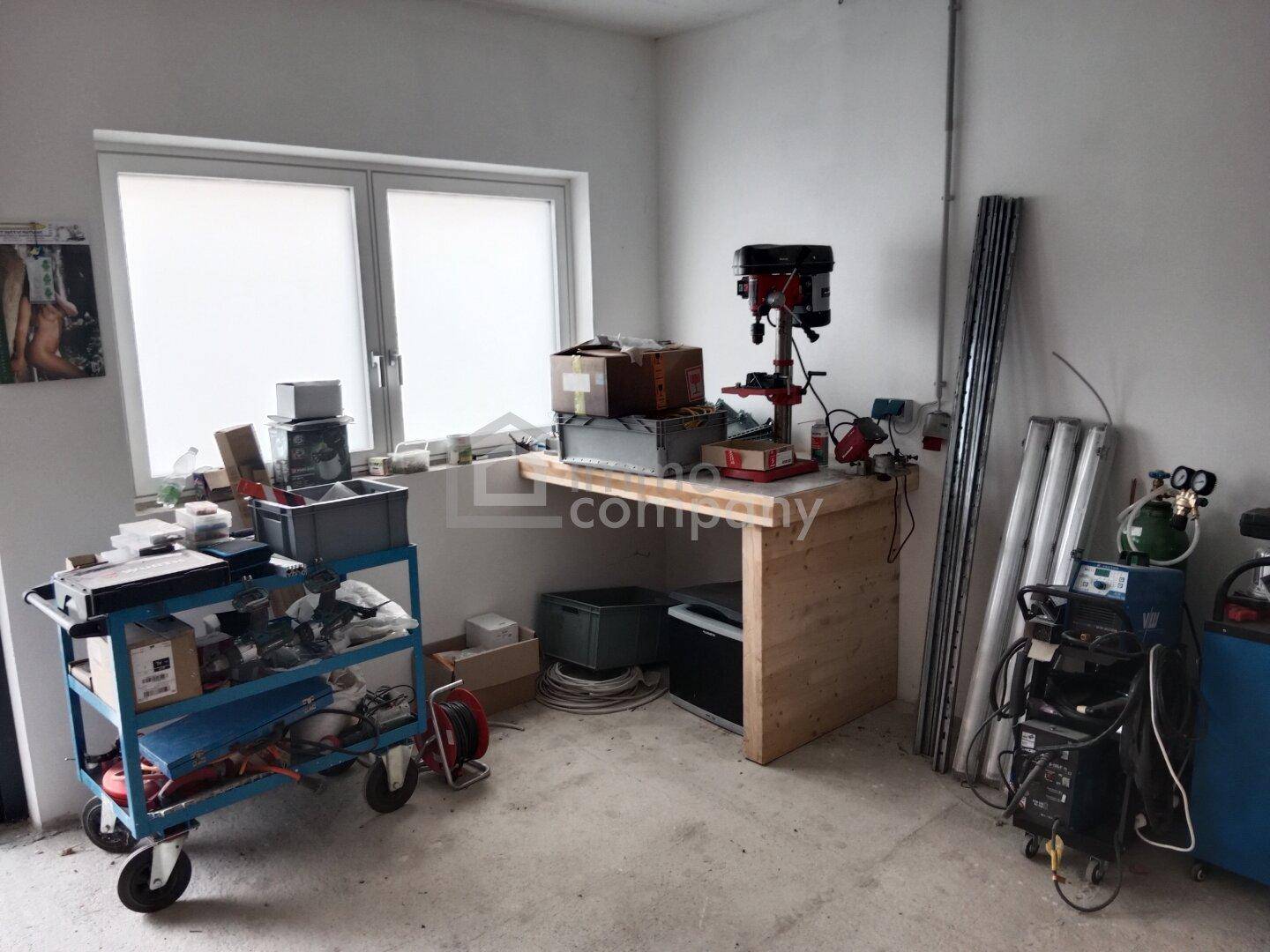 Heller Werkstattbereich mit Fenster, Bohrmaschine auf Werkbank und verschiedenen industriellen Geräten.