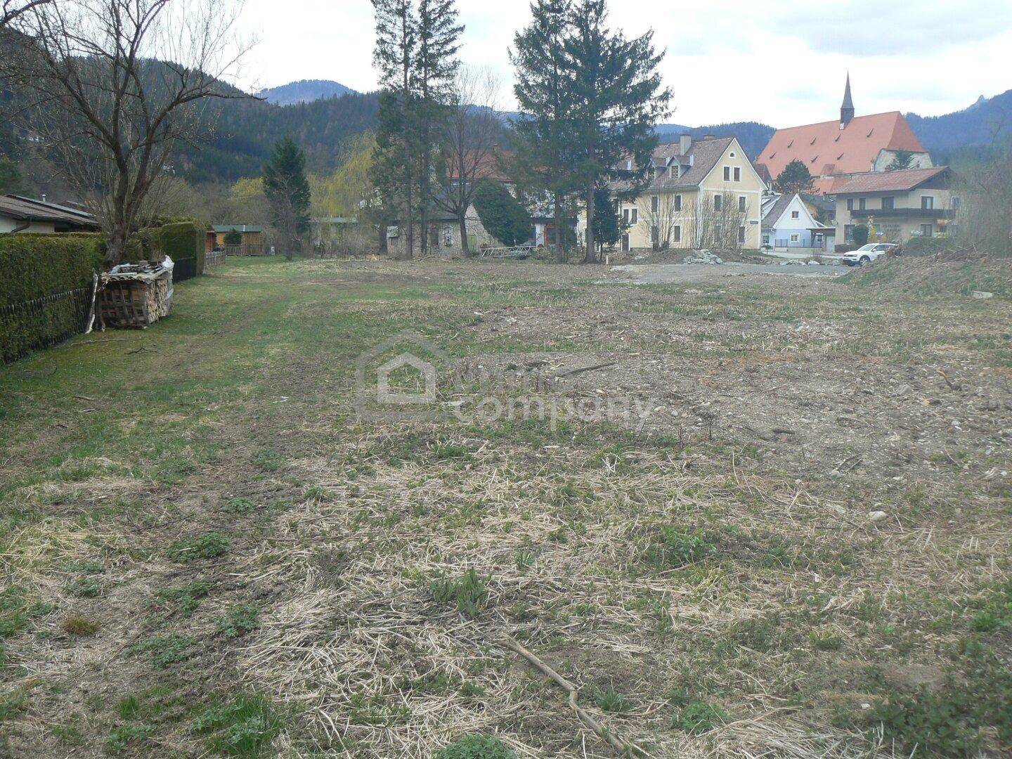 Unbebautes Grundstück mit Blick auf eine Hecke, Gebäude und eine Kirche im Hintergrund.