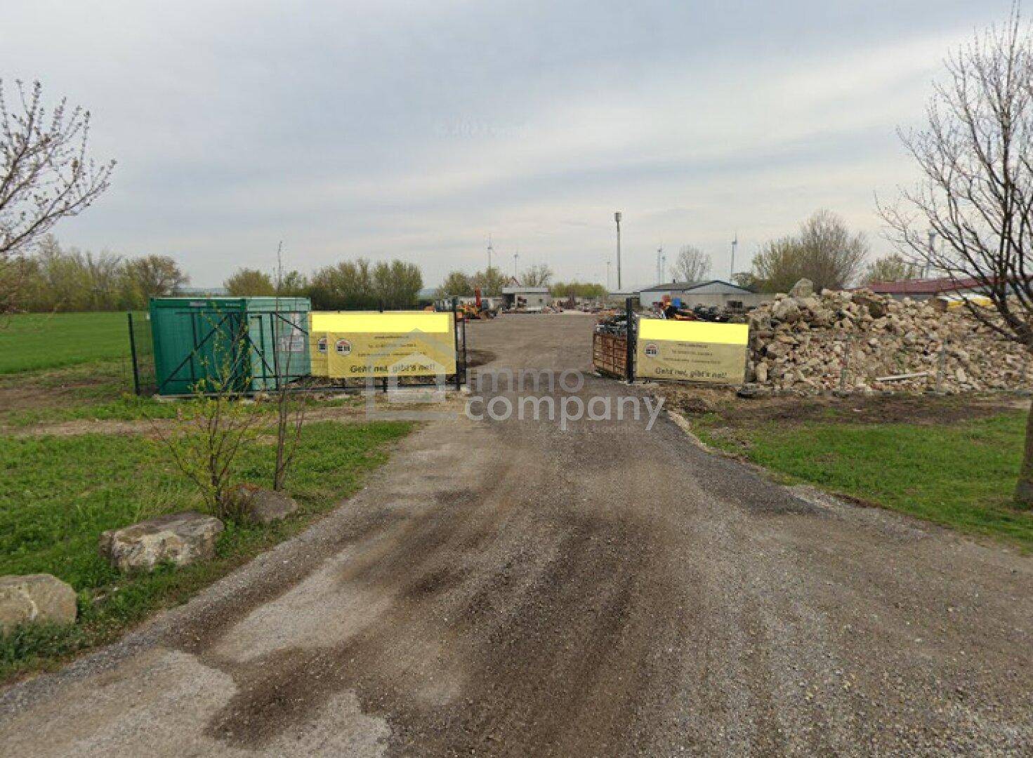 Schotterweg führt zu einem weitläufigen Gelände mit Bauschutt, Containern und Bäumen im Hintergrund.