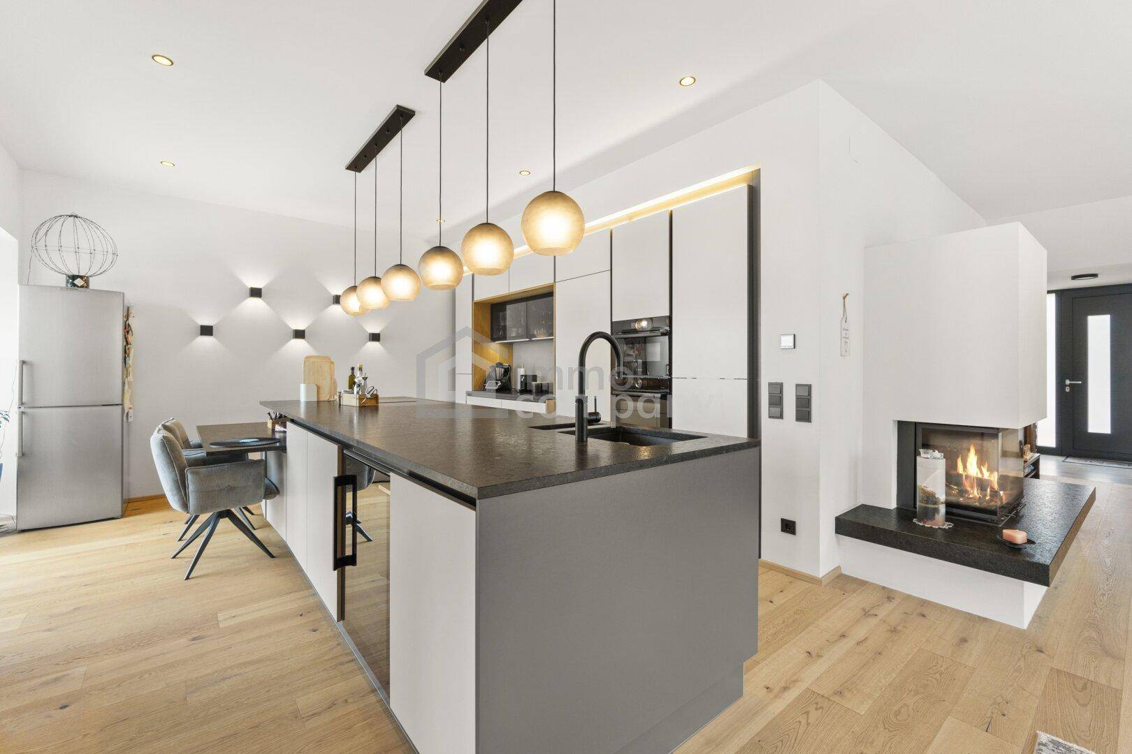 Moderne Küche mit Kochinsel, dunkler Arbeitsplatte, integrierten Geräten und Kamin im Hintergrund.