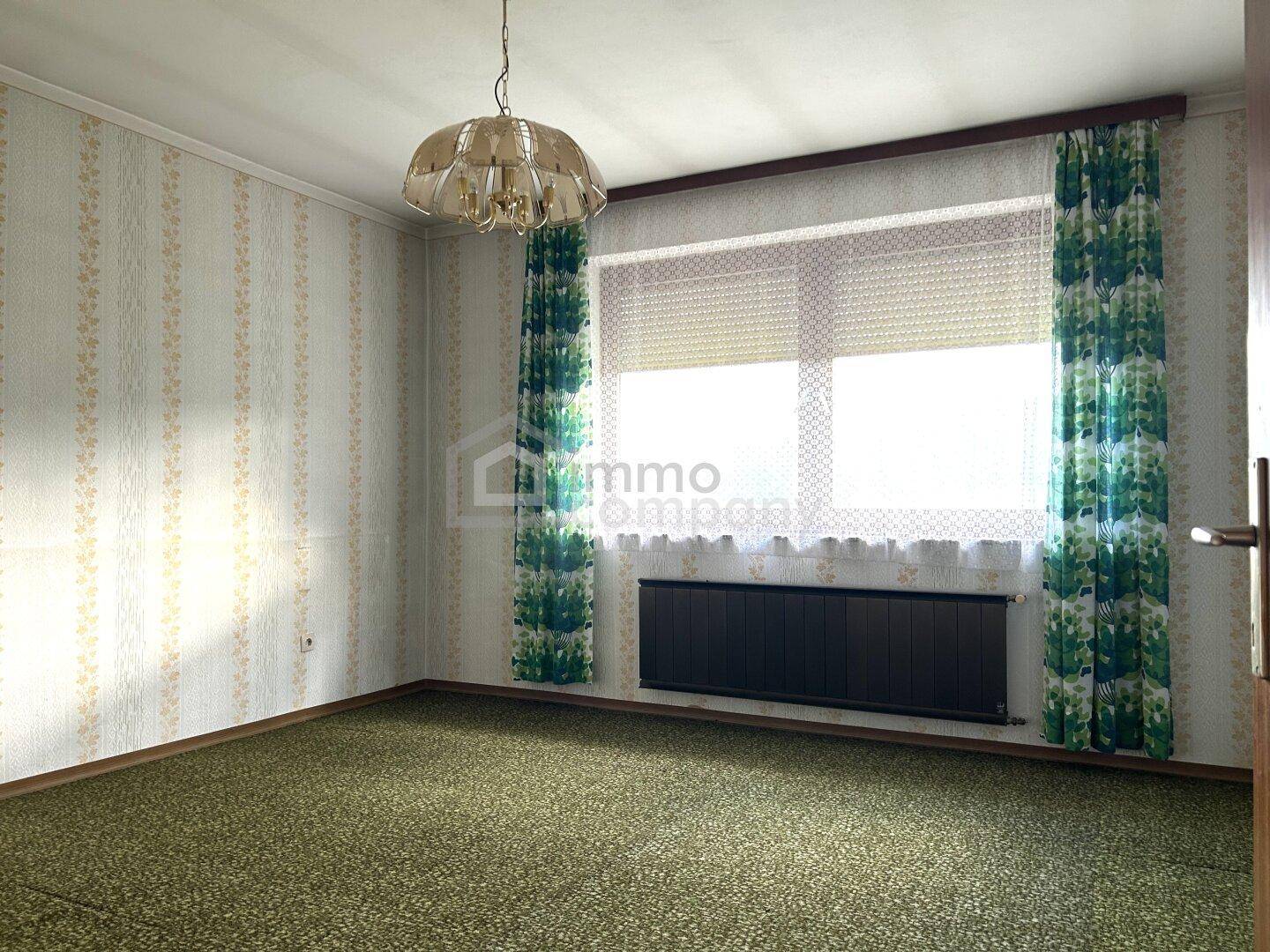 Schlafzimmer mit gemusterten Tapeten, Teppichboden und einem Fenster mit Blick ins Grüne.