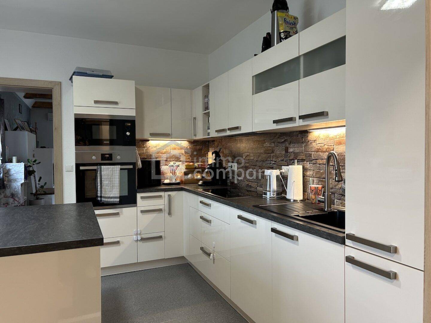 Moderne weiße Küche mit Hochglanzfronten, dunkler Arbeitsplatte und Backstein-Spritzschutz.