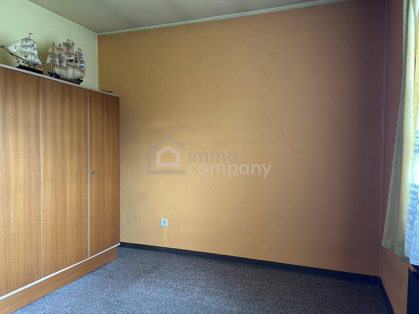 Schlafzimmer mit schlichten Wänden, Teppichboden und einem großen Holzschrank.