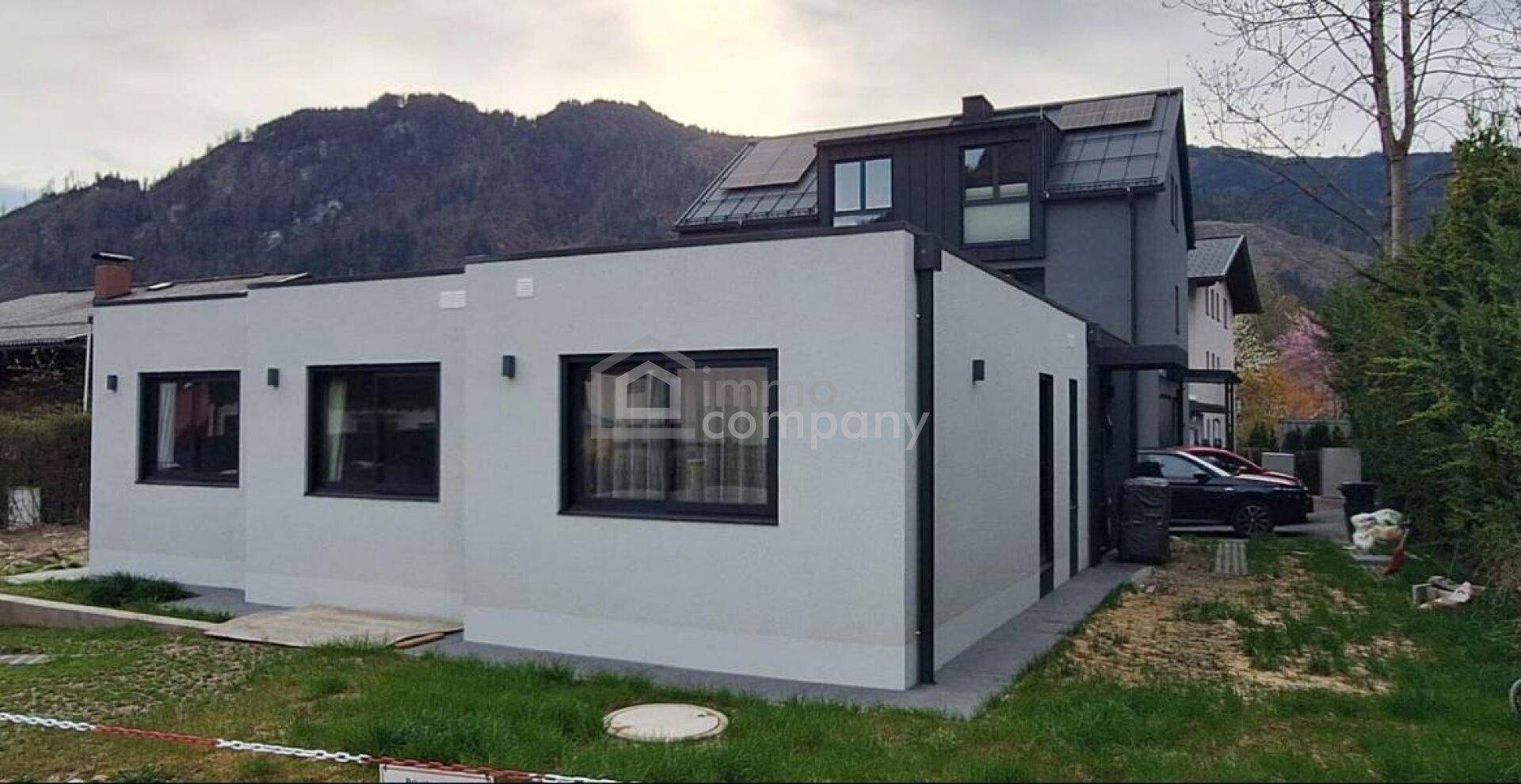 Ansicht des modernen Hauses mit dunklem Dach und Fenstern, umgeben von Natur und Bergen.