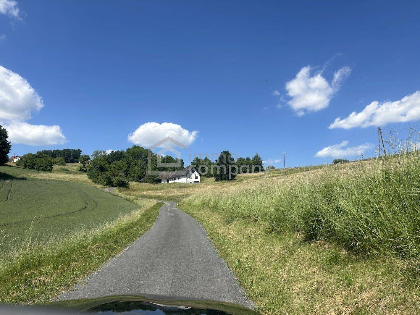 Weitläufige ländliche Landschaft mit einer Straße, die zu einem entfernten Haus führt, umgeben von Feldern.