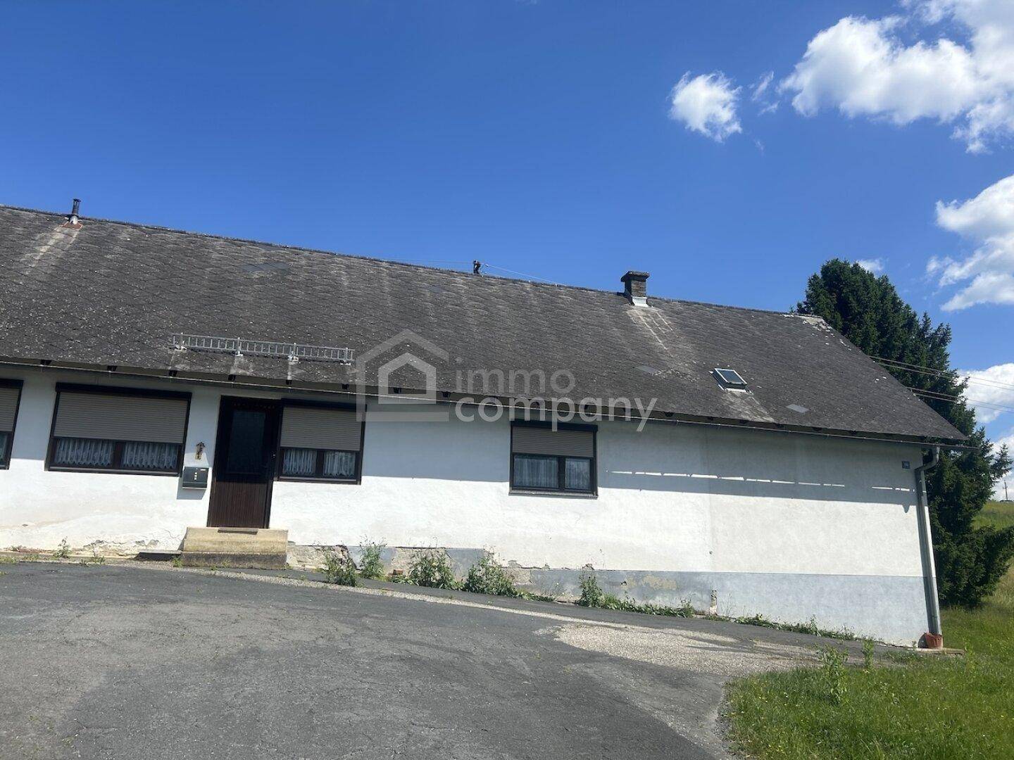 Fassade eines älteren Gebäudes mit weißem Putz, dunklem Dach und Fenstern mit Rollläden.