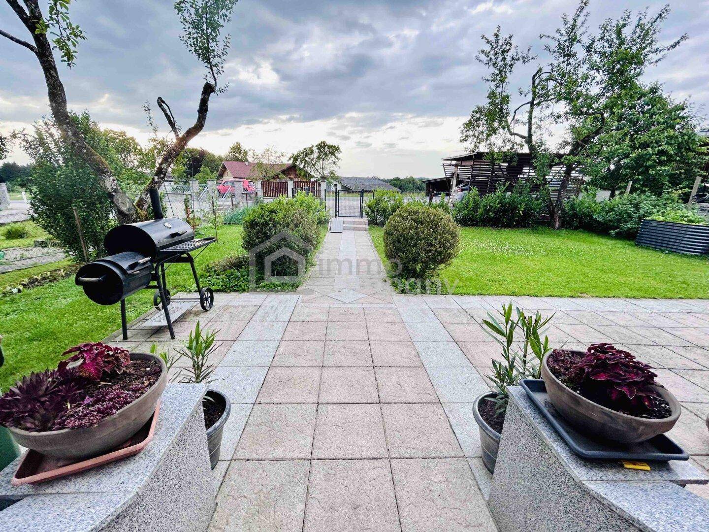 Gepflasterte Terrasse mit Grillbereich und Zugang zu einem weitläufigen Garten unter bewölktem Himmel.