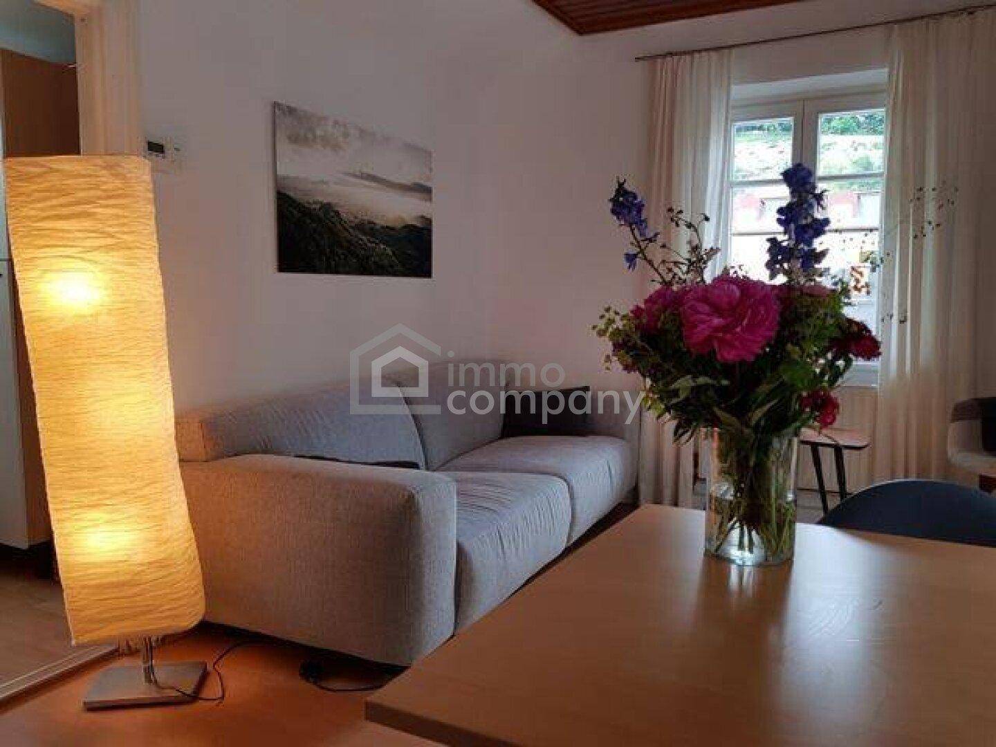 Helles Wohnzimmer mit beigem Sofa, Holztisch und Blumenstrauß, durchflutet von natürlichem Licht.