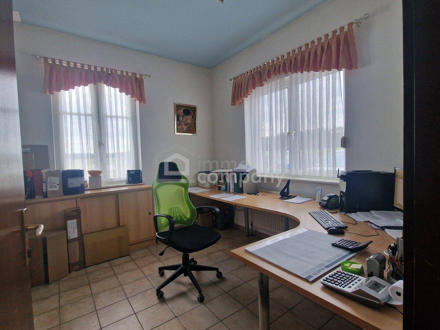 Kleines Büro mit Schreibtisch, Bürostuhl und zwei Fenstern, die viel Tageslicht hereinlassen.