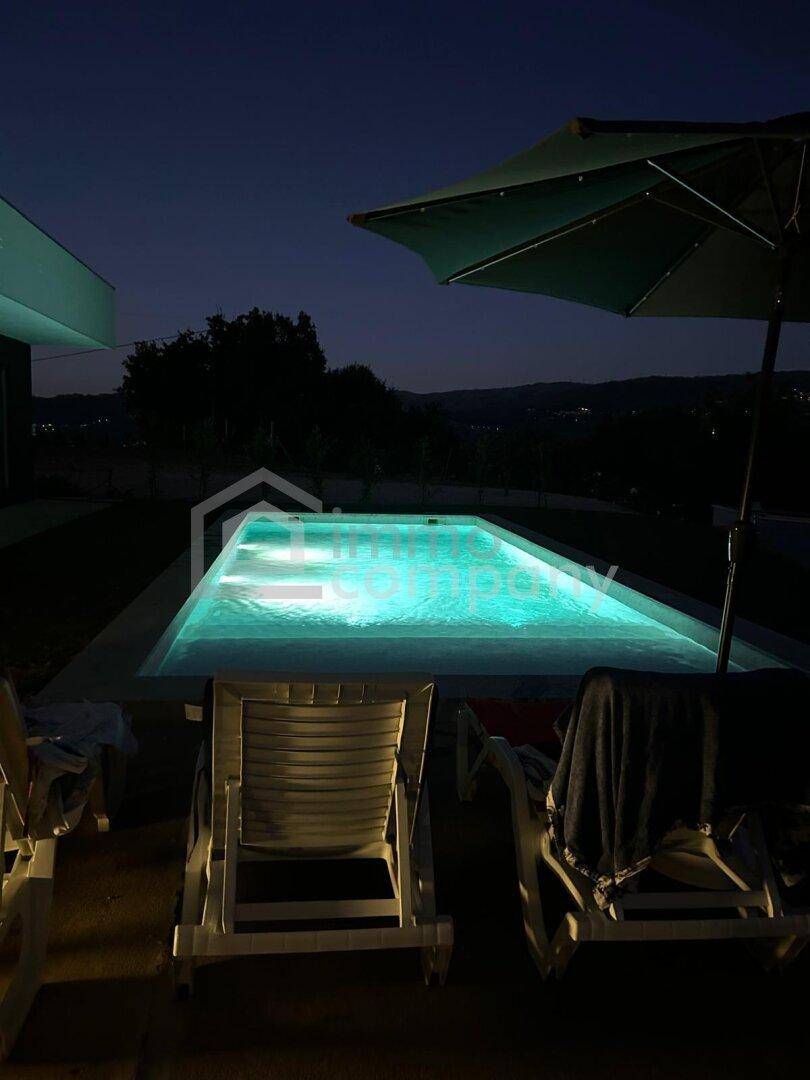 Beleuchteter Swimmingpool bei Nacht mit Liegestühlen und Schirm, umgeben von Dunkelheit.