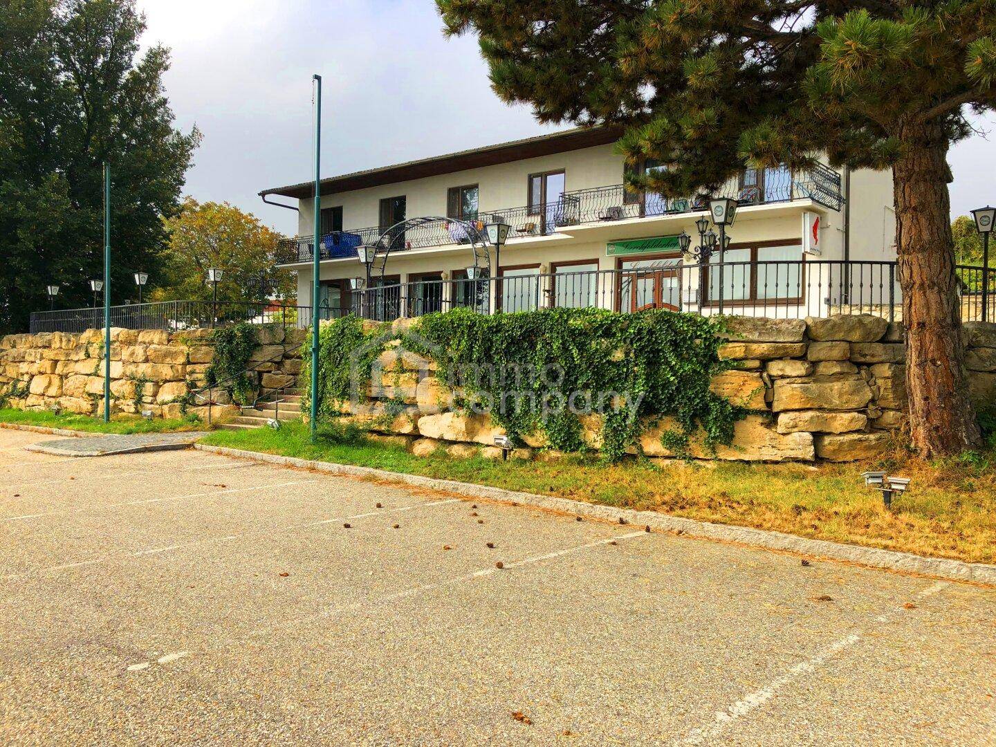 Gebäudefassade mit Balkonen und einem vorgelagerten Parkplatz, umgeben von einer Natursteinmauer.