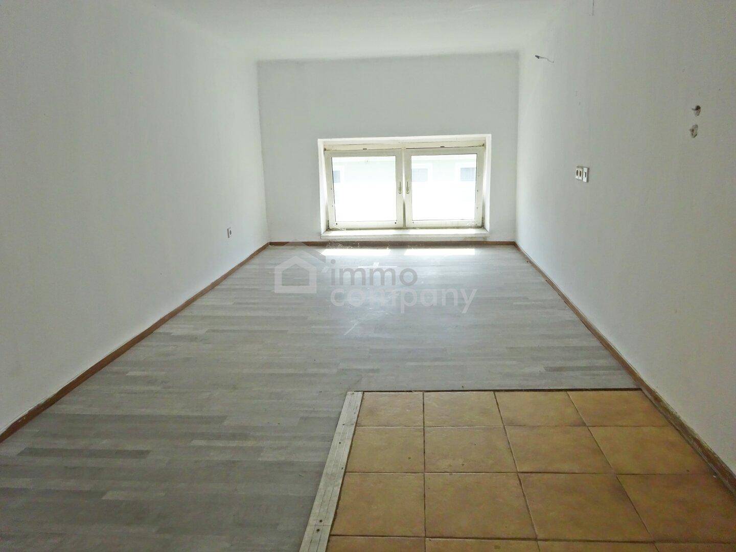 Heller, leerer Raum mit Fenster, weißen Wänden und einer Kombination aus Laminat- und Fliesenboden.