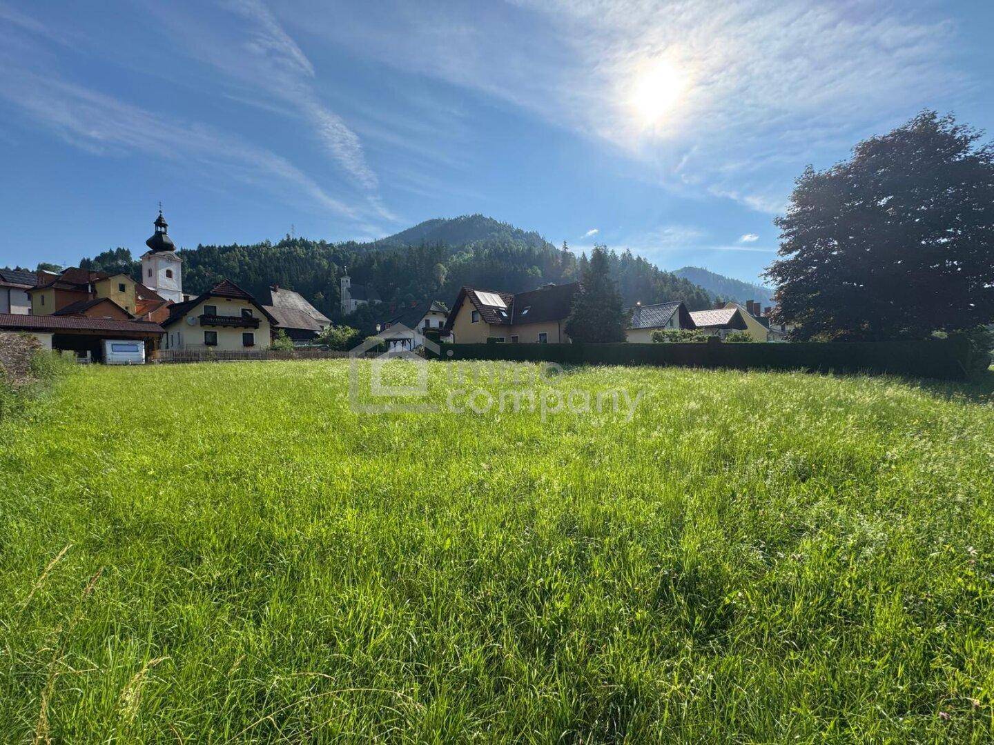 Sonniges, weitläufiges Grundstück mit Blick auf die Berge und die Dorfkirche unter blauem Himmel.