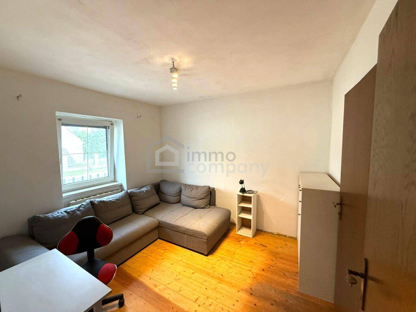 Helles Wohnzimmer mit Ecksofa, Holzboden und einem Fenster mit Blick ins Grüne.