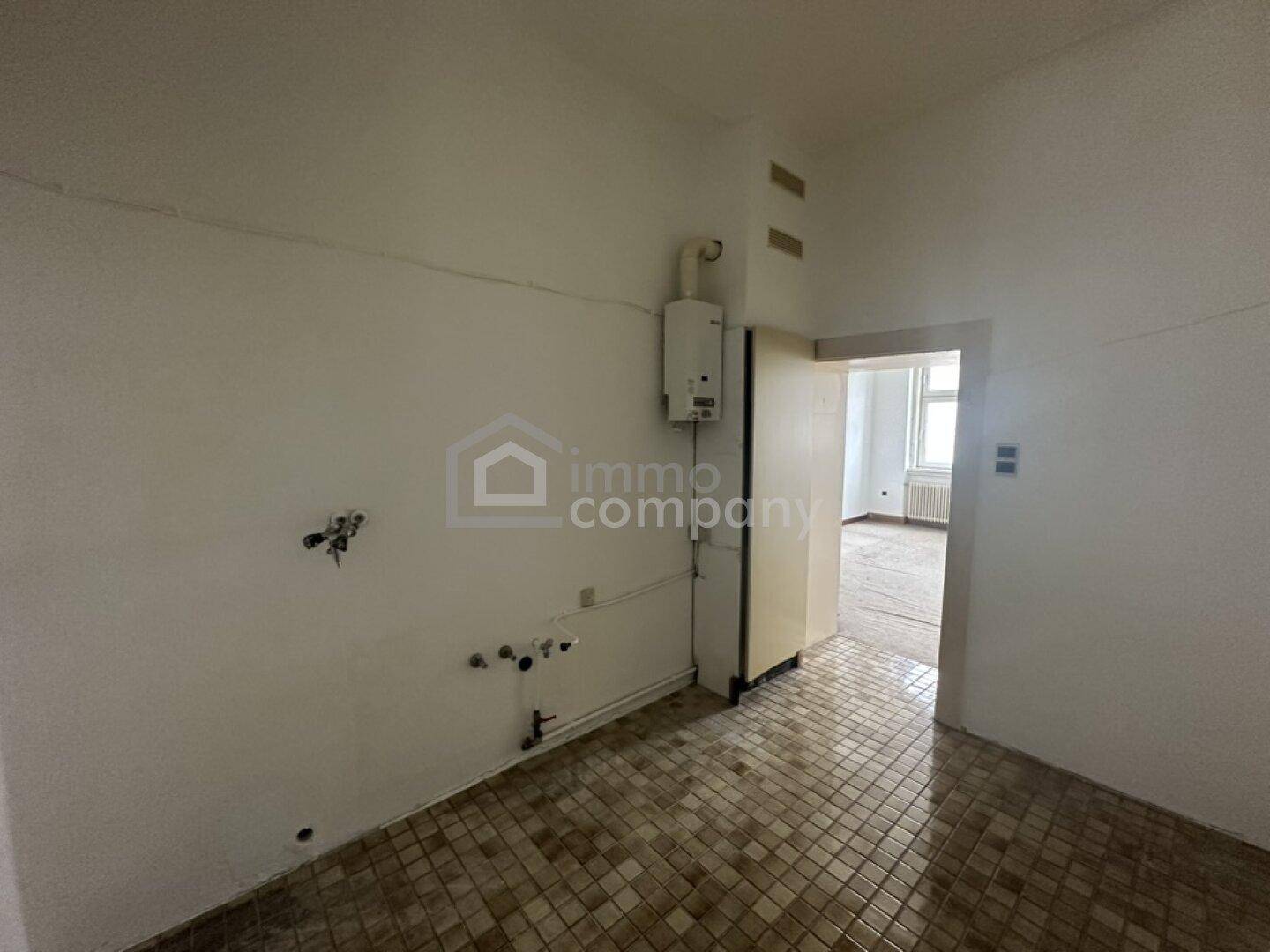 Leerer Küchenraum mit Fliesenboden, Wasseranschlüssen und Durchgang zu einem weiteren Zimmer.