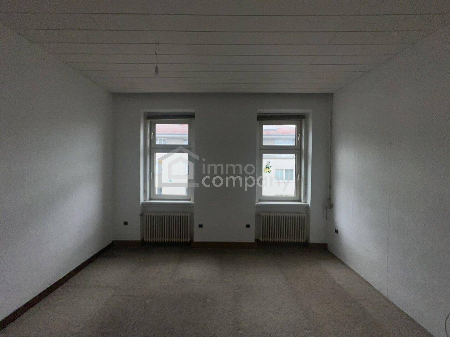 Helles Zimmer mit zwei großen Fenstern und Heizkörpern, bereit für Renovierungsarbeiten.