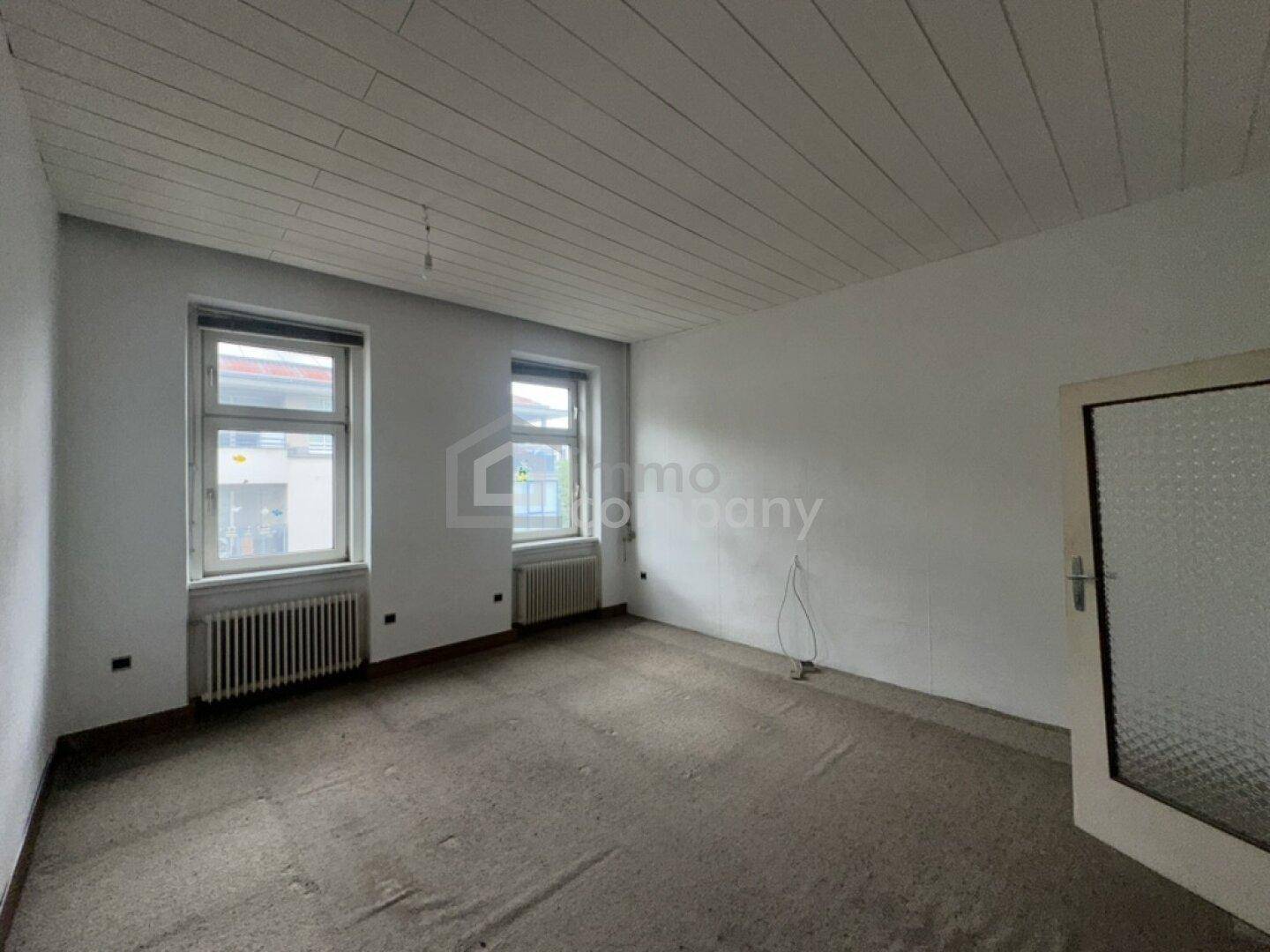 Heller, unmöblierter Raum mit zwei Fenstern, Heizkörpern und einer Holzpaneeldecke.