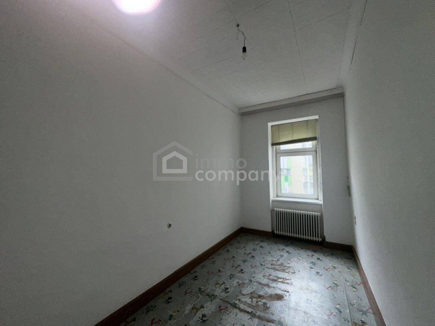 Kleiner, heller Raum mit einem Fenster, Heizkörper und altem gemustertem Bodenbelag.