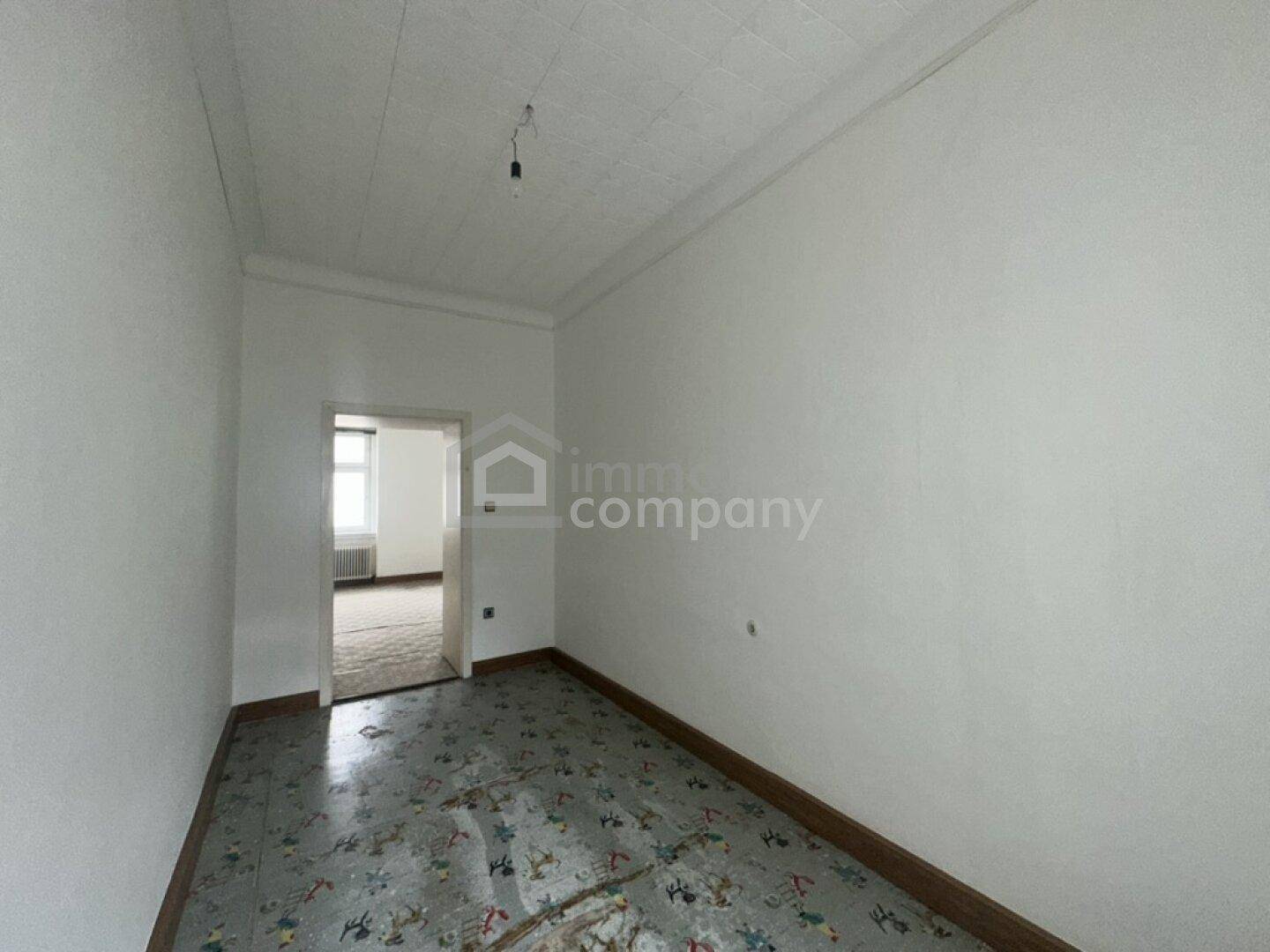 Heller Raum mit Durchgang zu einem weiteren Zimmer und altem gemustertem Bodenbelag.