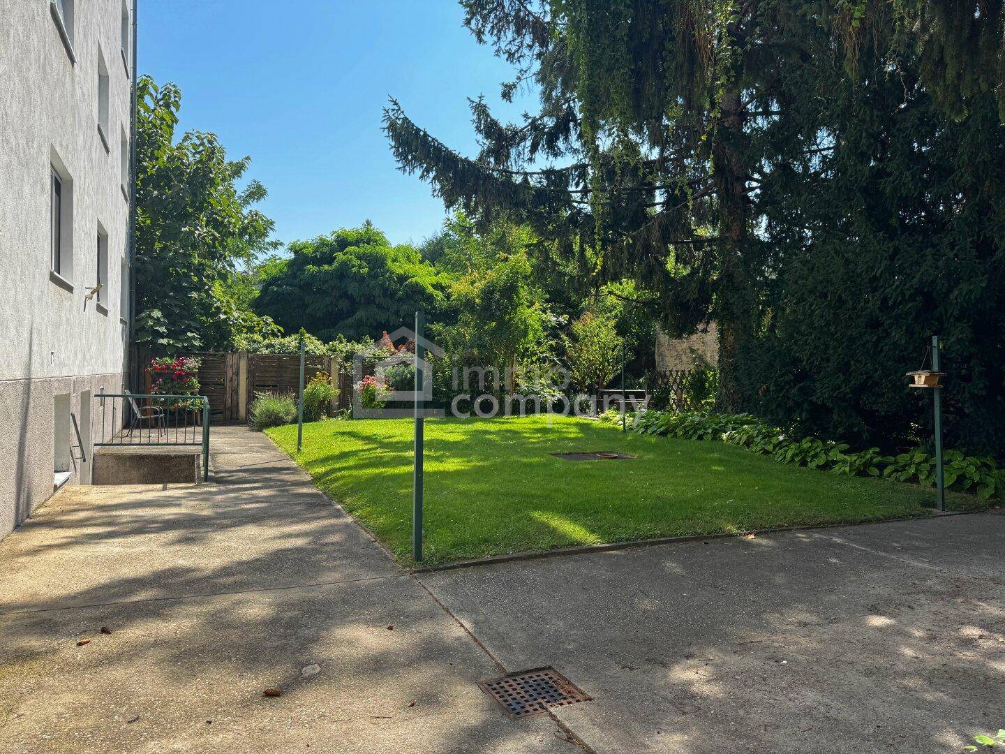 Gepflegter Gartenbereich mit Rasenfläche, Bäumen und einem gepflasterten Weg neben dem Gebäude.