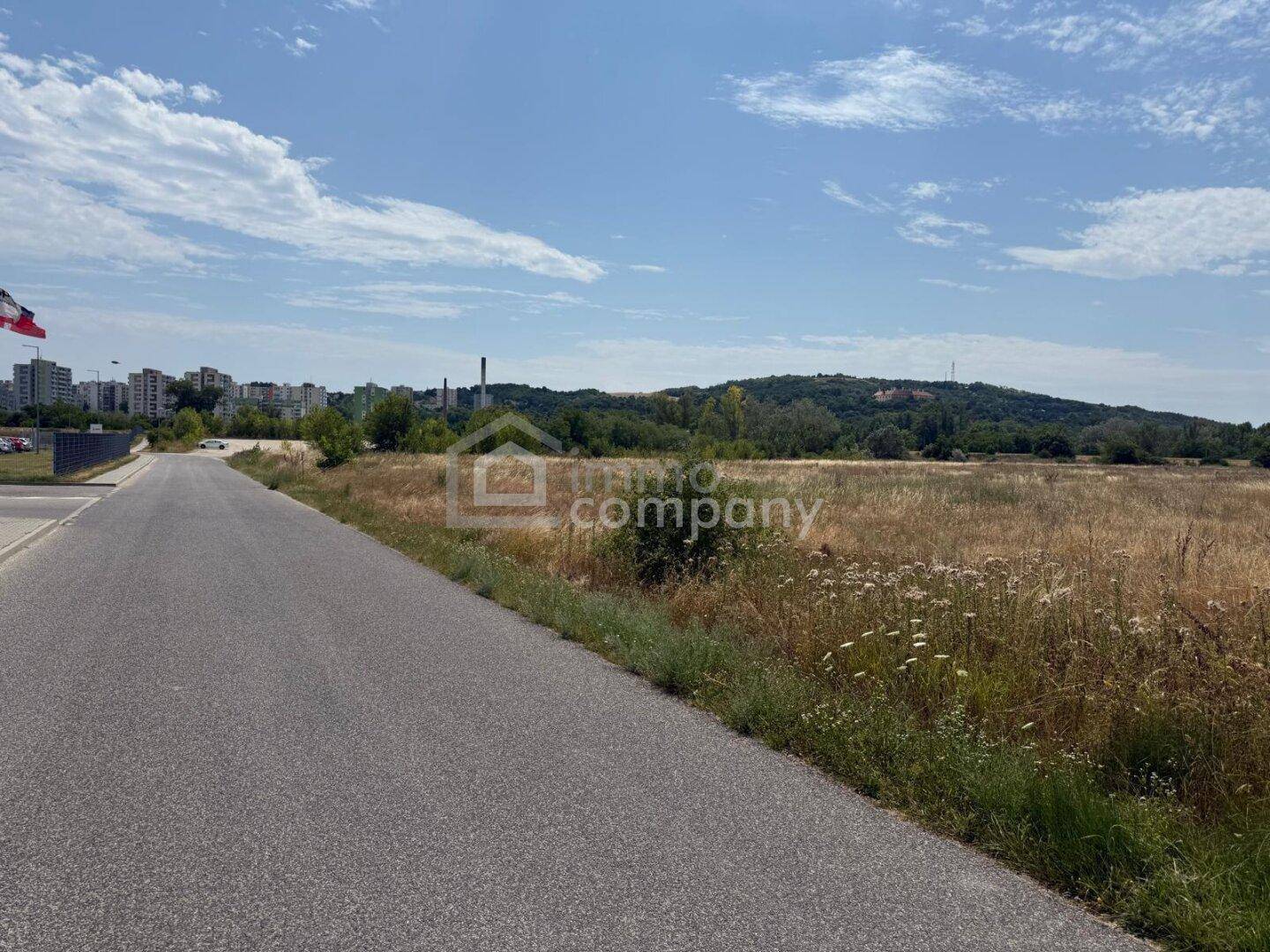 Weitläufiges unbebautes Land neben einer Straße mit Blick auf entfernte Gebäude und Hügel.