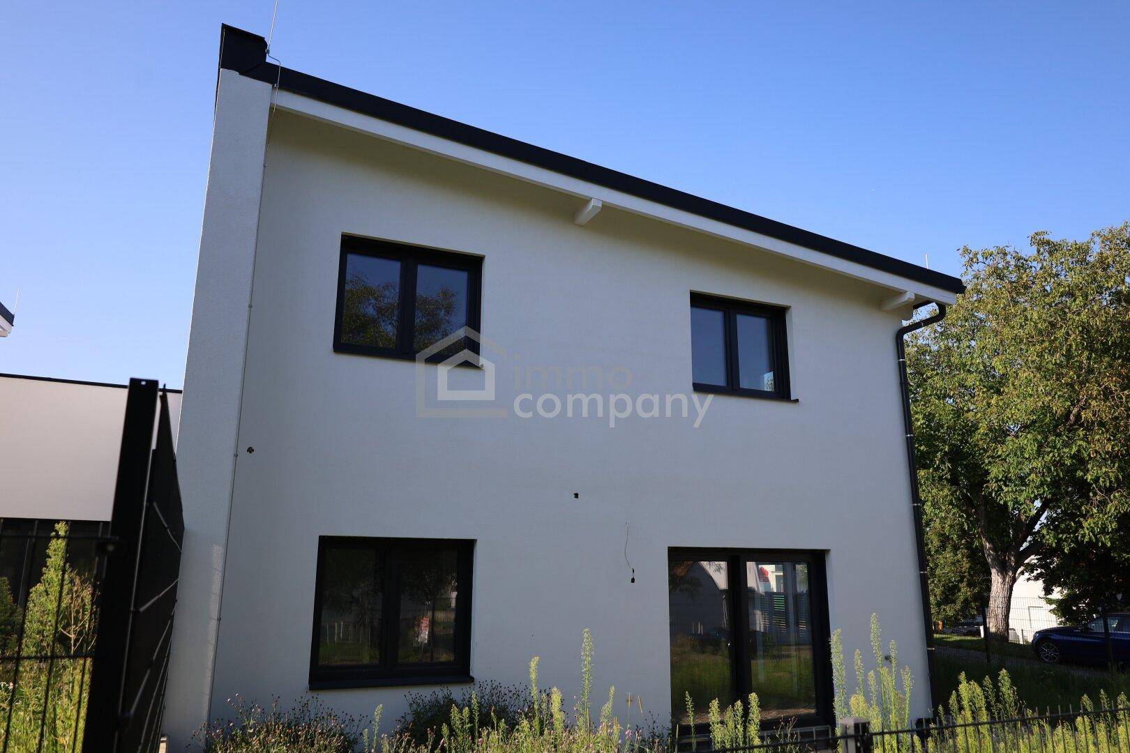 Moderne Hausfassade in Weiß mit dunklen Fensterrahmen und klaren Linien unter blauem Himmel.