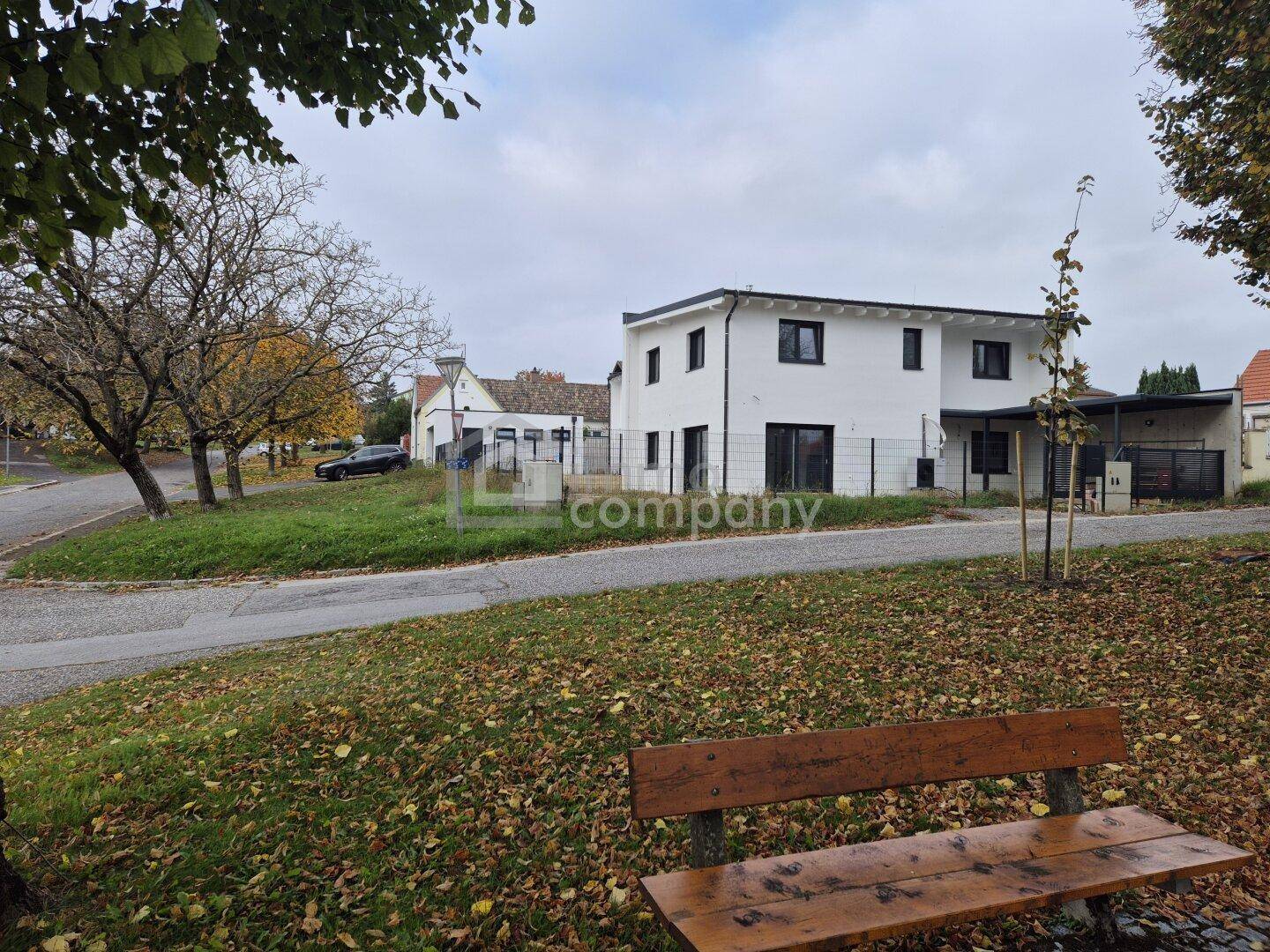 Weißes Neubauhaus in ländlicher Umgebung mit Herbstlaub und einer Parkbank im Vordergrund.