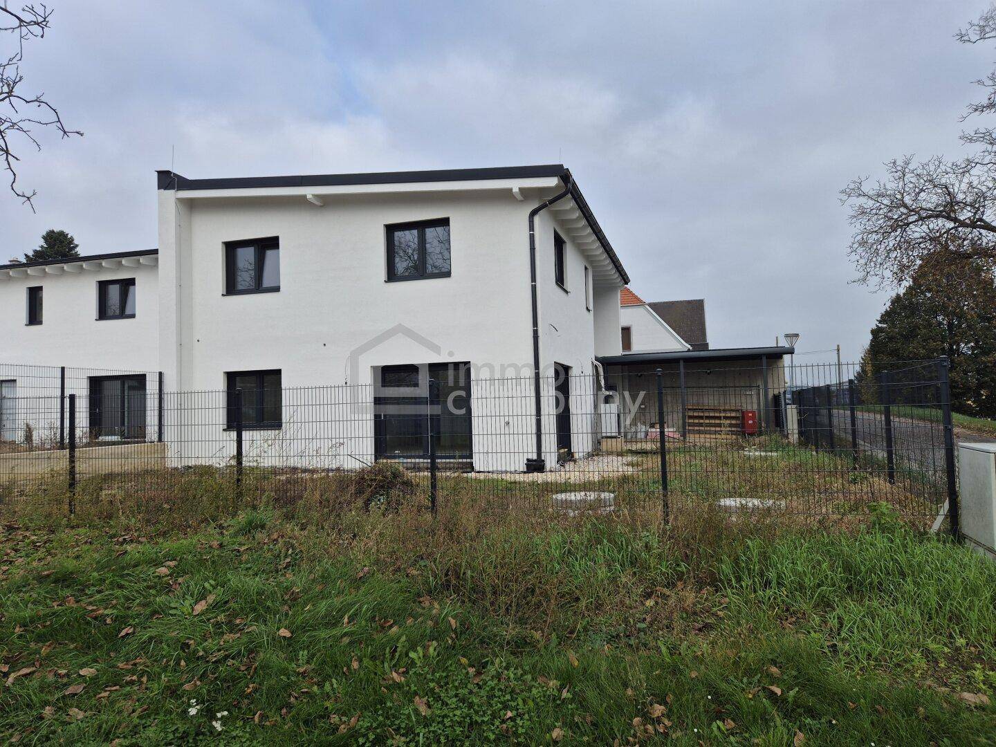Rückansicht eines modernen Neubaus mit Gartenbereich, Zaun und einem Carport im Hintergrund.