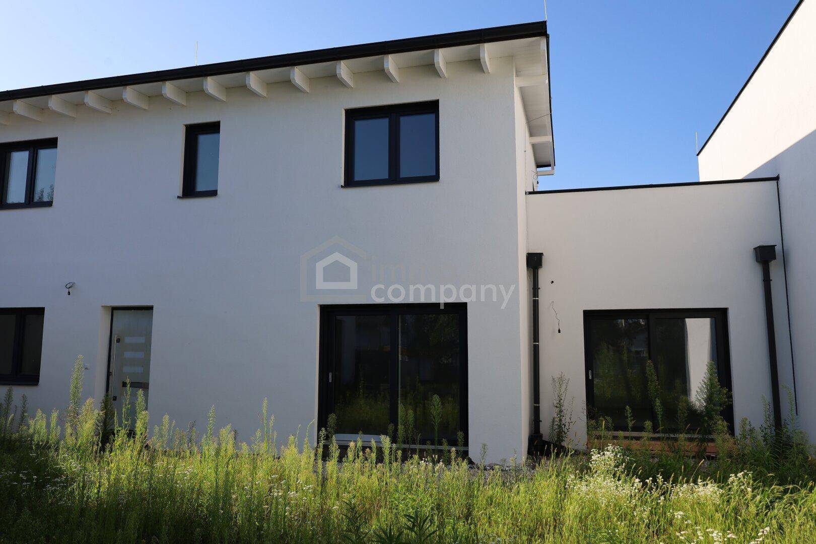Rückseite eines modernen weißen Hauses mit schwarzen Fensterrahmen und Zugang zum noch ungestalteten Gartenbereich.