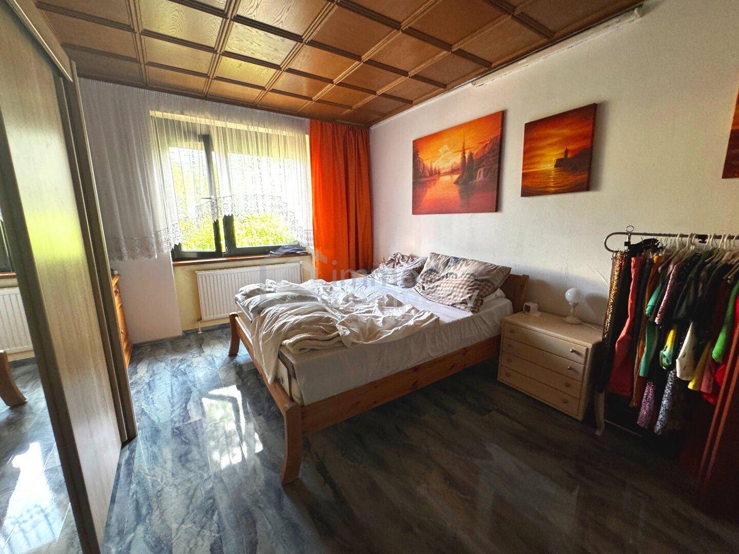 Schlafzimmer mit Doppelbett, Holzdecke, Fliesenboden und Blick ins Grüne durch das Fenster.
