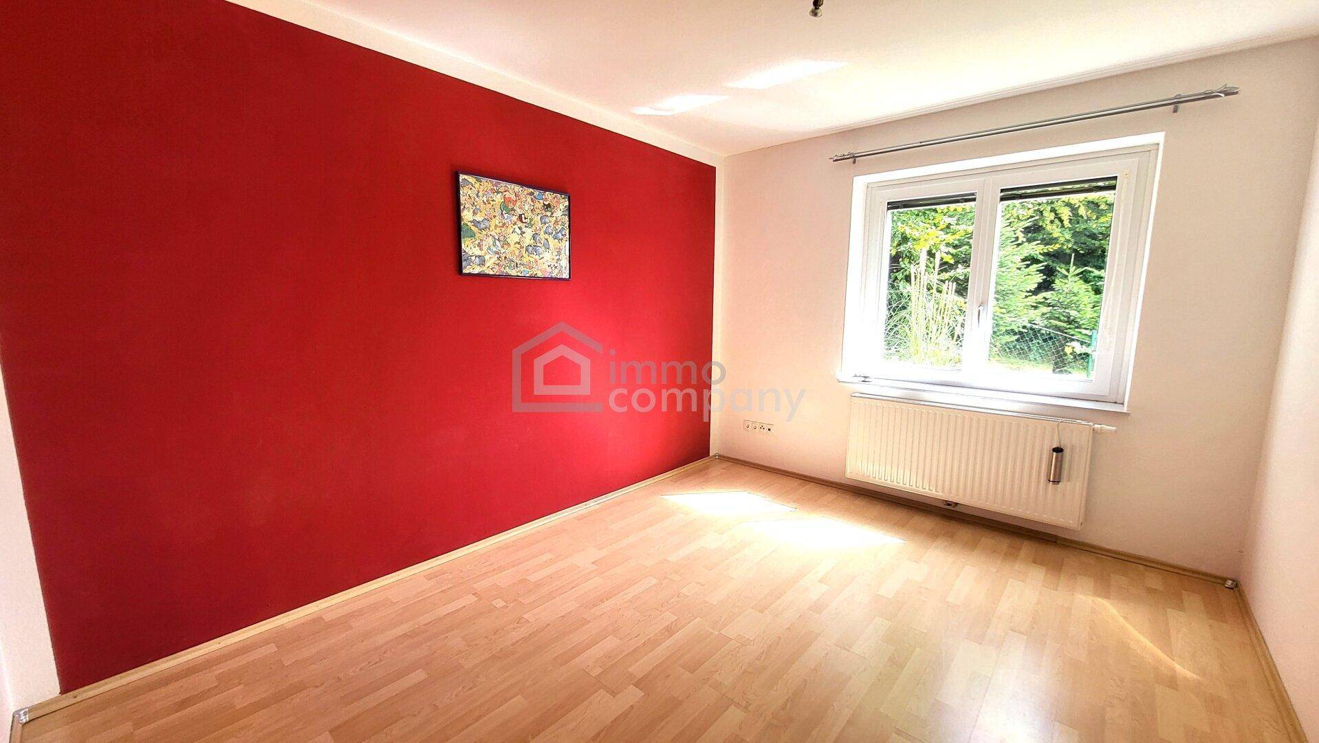 Leeres Zimmer mit roter Akzentwand, Laminatboden und großem Fenster mit Blick ins Grüne.