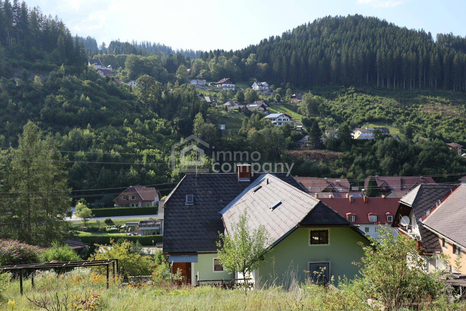 Panoramablick auf eine Siedlung in einer hügeligen, bewaldeten Landschaft unter blauem Himmel.