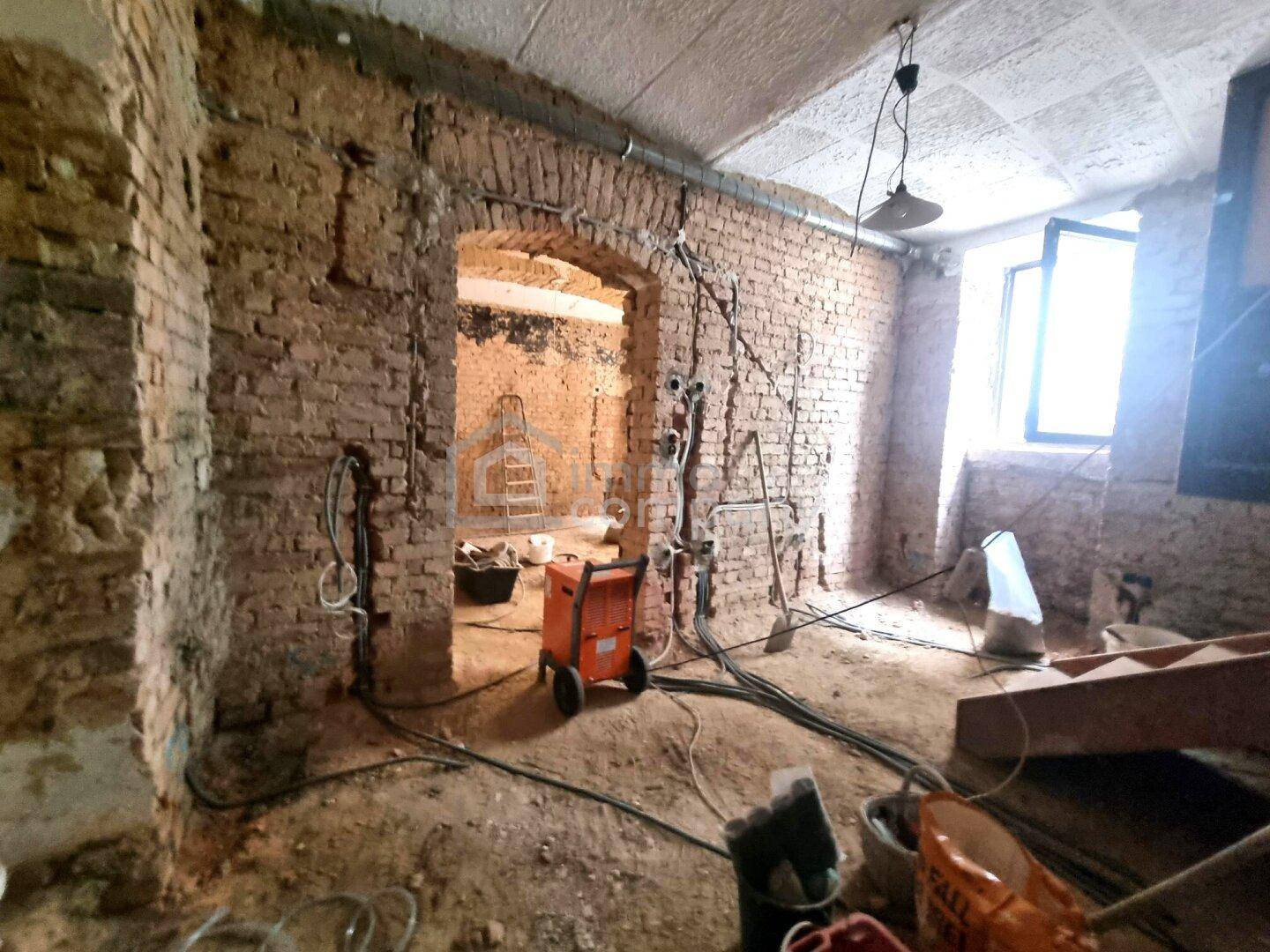 Innenraum im Rohzustand mit Ziegelwänden, Baugeräten und natürlichem Lichteinfall durch ein Fenster.