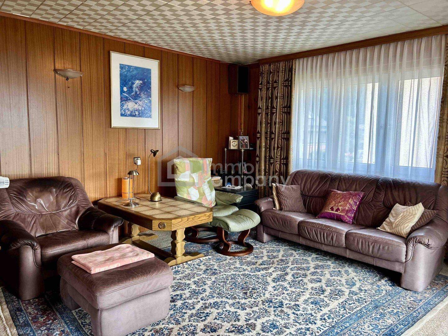Gemütliches Wohnzimmer mit dunklen Ledersofas, Holzwänden und großem Fenster.