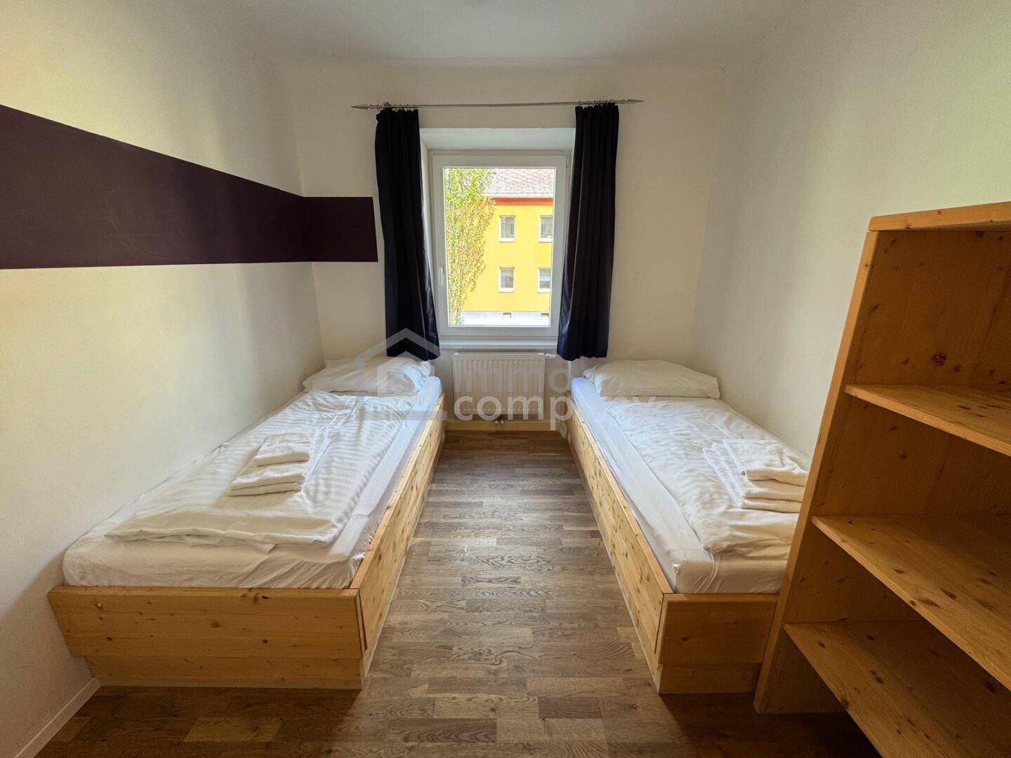 Helles Schlafzimmer mit zwei Einzelbetten, Holzregal und Blick ins Grüne.