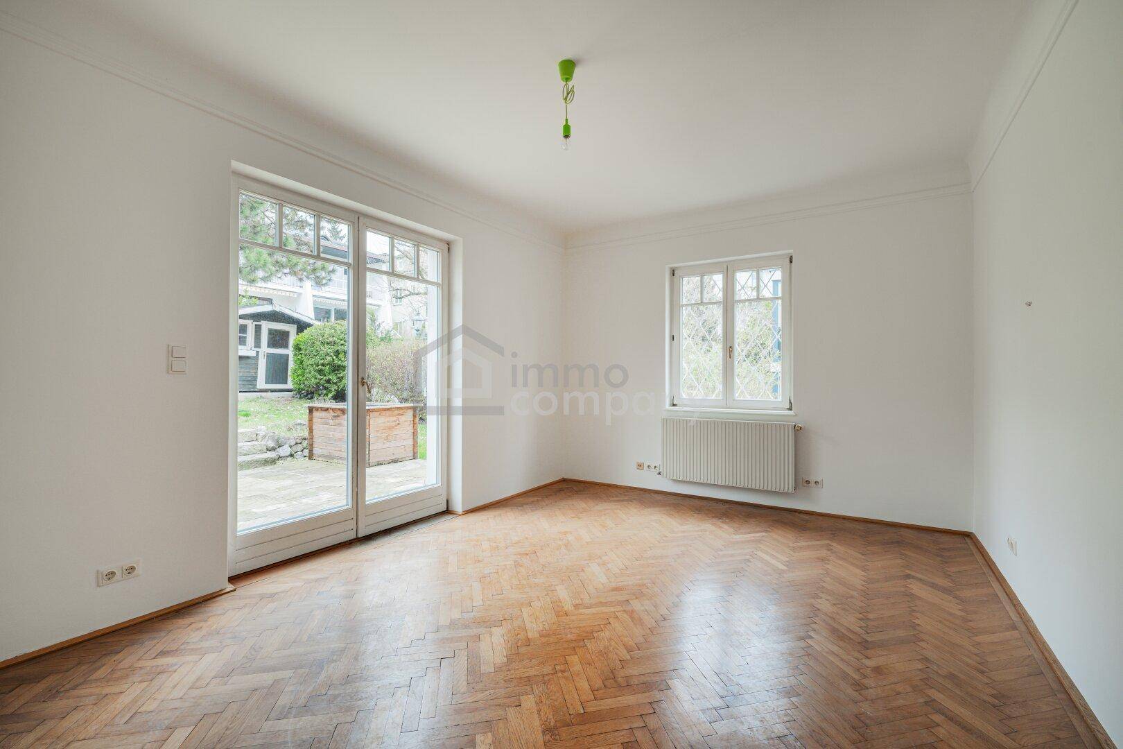 Heller Raum mit Fischgrätparkett und großen Fenstertüren, die Zugang zum Garten bieten.