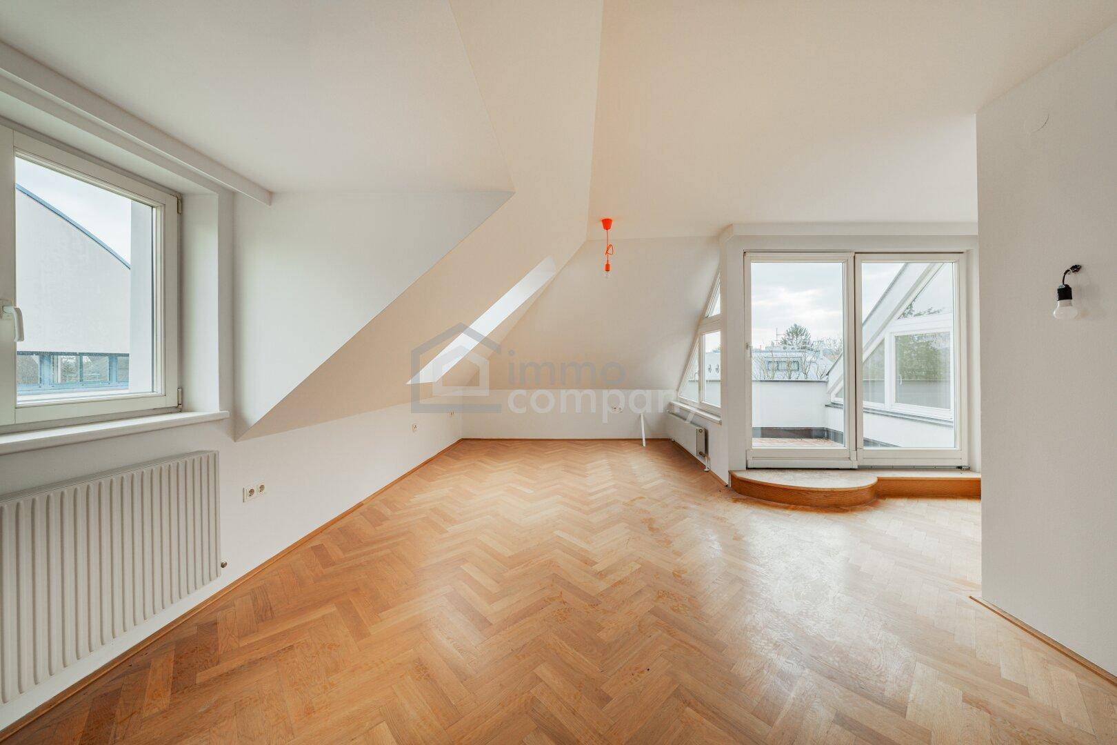 Geräumiger Dachgeschossraum mit Fischgrätparkett, schrägen Wänden und Zugang zu einer Dachterrasse.