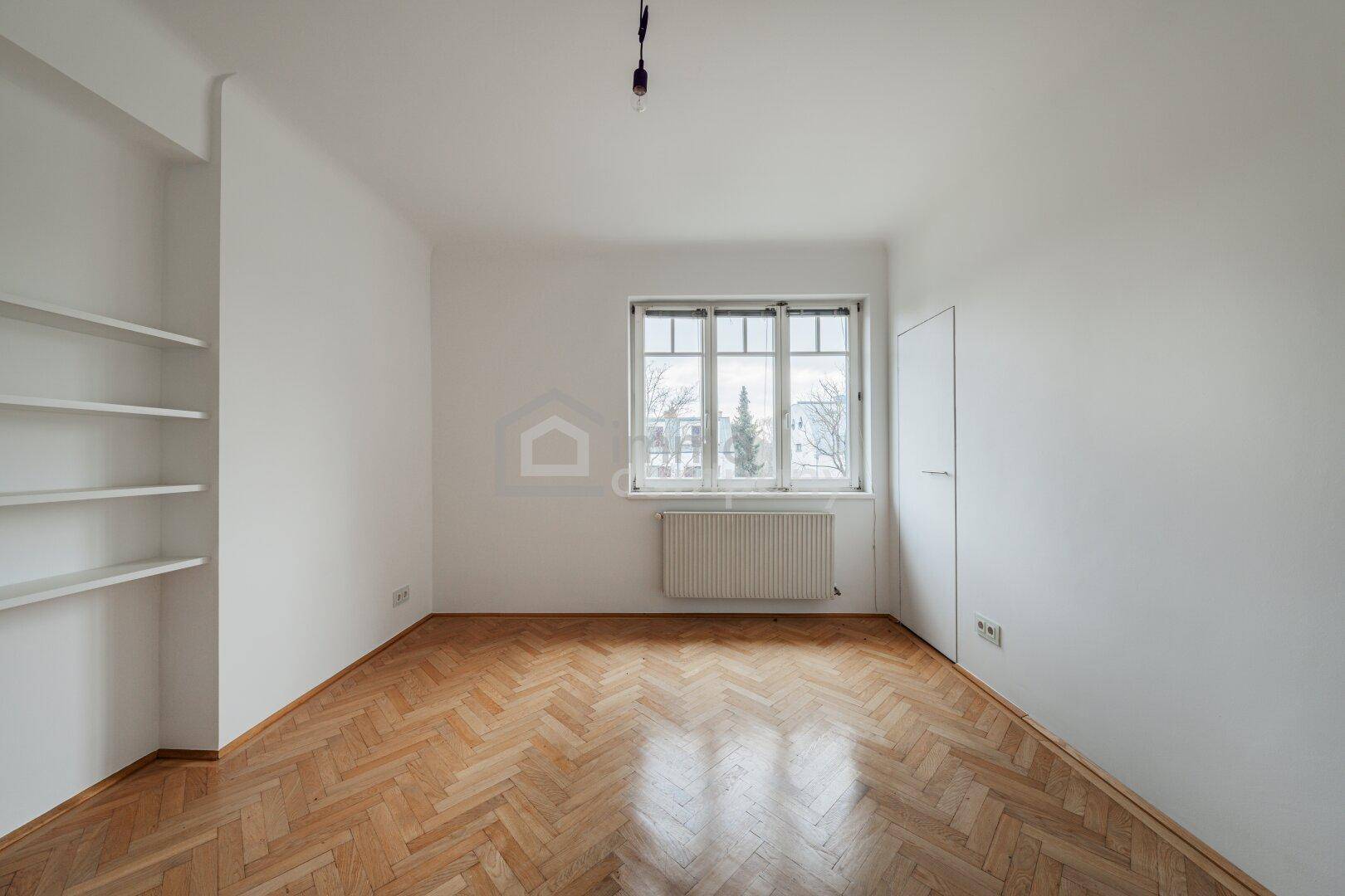 Heller Raum mit Fischgrätparkett, Einbauregalen und großem Fenster, ideal als Schlaf- oder Arbeitszimmer.