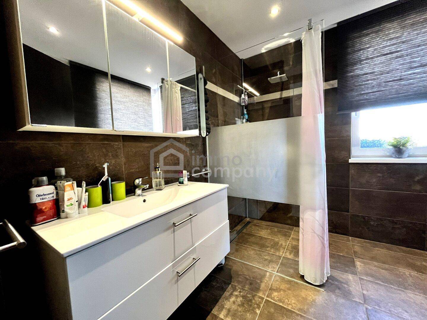 Modernes Badezimmer mit dunklen Fliesen, Duschbereich und Waschtisch mit Spiegelschrank und Fenster.