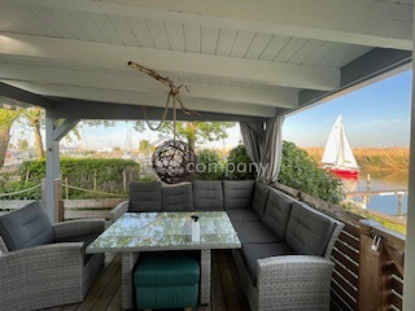 Überdachte Terrasse mit bequemen Outdoor-Möbeln und direktem Blick auf das Wasser mit Segelboot.
