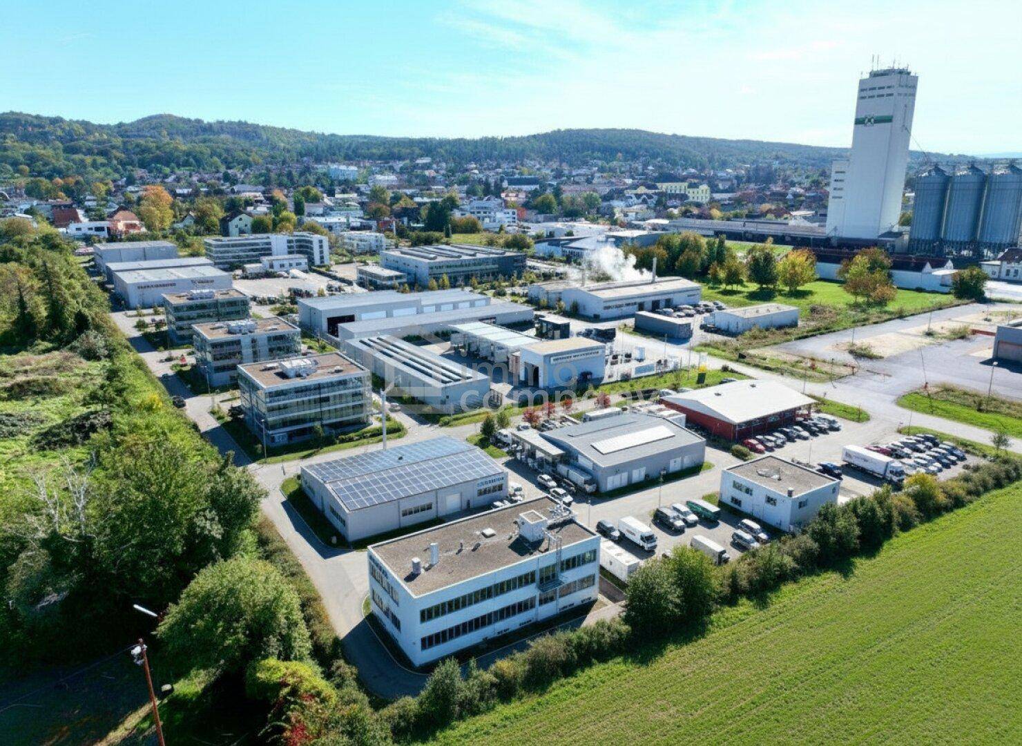 Luftaufnahme eines Industriegebiets mit verschiedenen Gewerbegebäuden, Solaranlagen und einem großen Silo im Hintergrund.