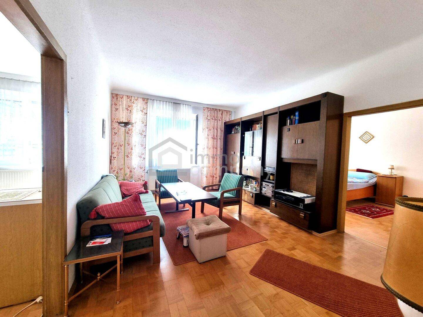 Traditionell eingerichtetes Wohnzimmer mit Holzmöbeln, Teppichen und Blick in ein Schlafzimmer.