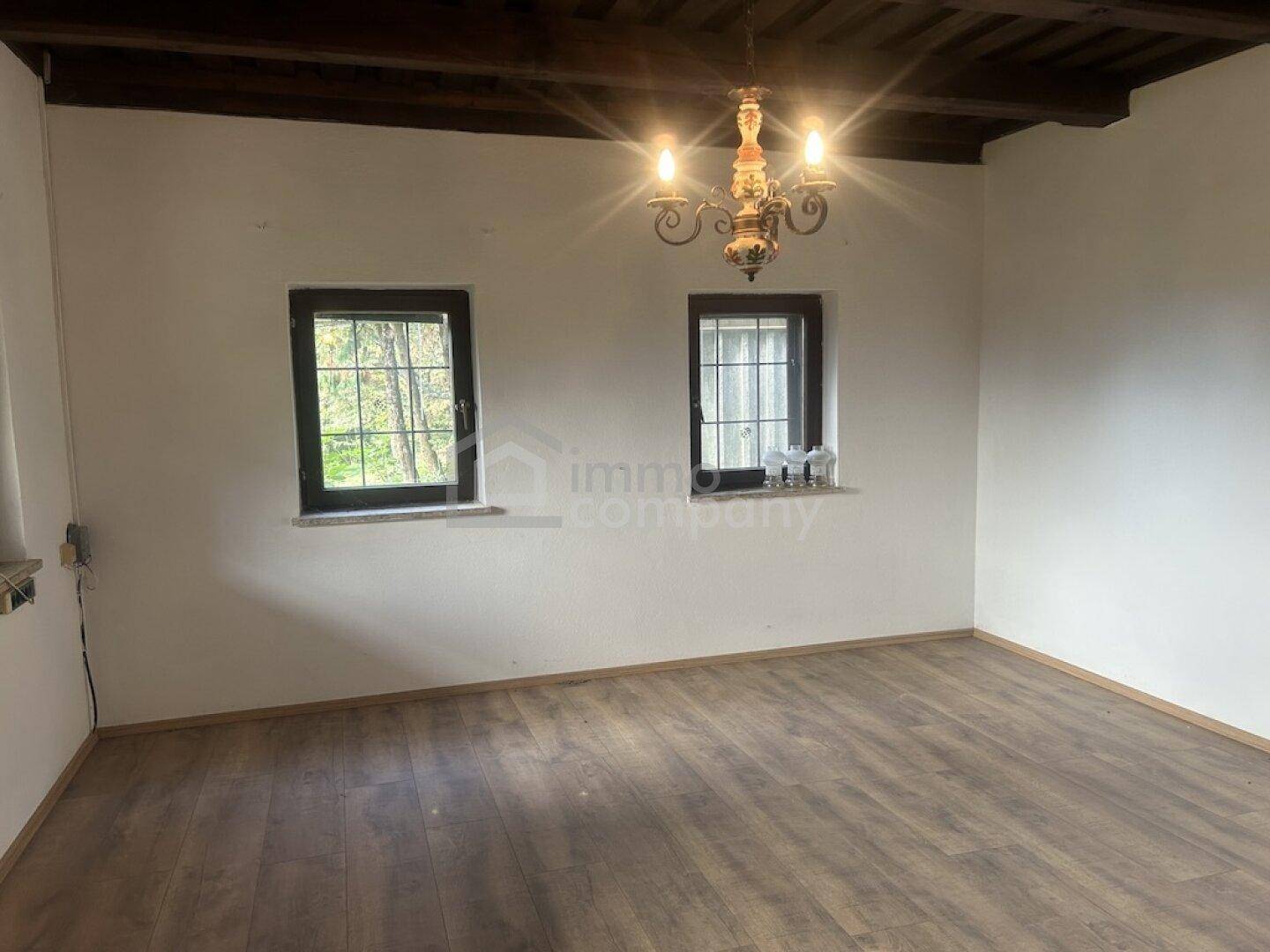 Heller Wohnbereich mit zwei Fenstern, Laminatboden und einer dekorativen Deckenleuchte.