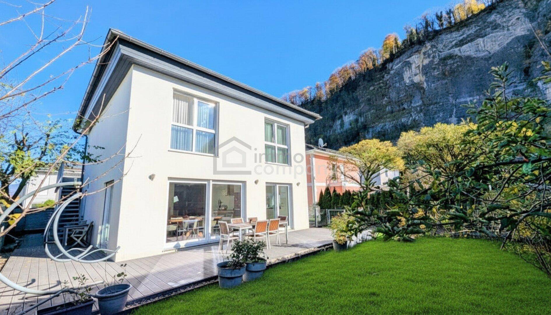 Modernes Einfamilienhaus mit großer Terrasse und Garten, umgeben von einer Felswand und viel Grün.
