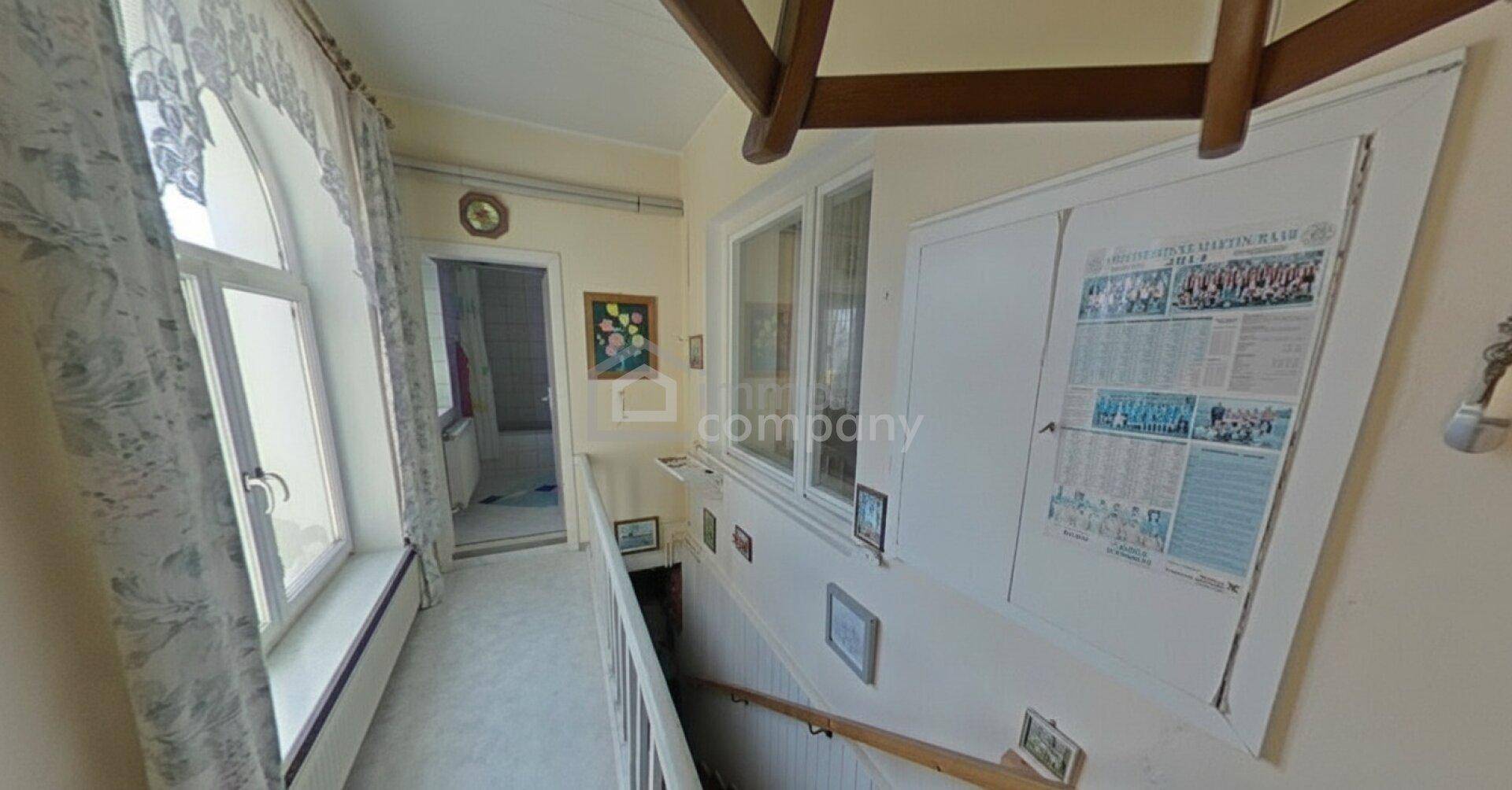 Helles Treppenhaus mit großen Fenstern, Holzgeländer und dekorativen Elementen.