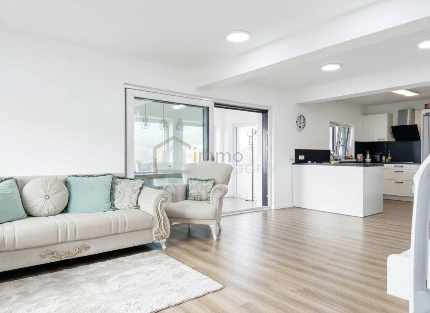 Lichtdurchfluteter Wohnbereich mit beigem Sofa, Laminatboden und Blick in die offene Küche.